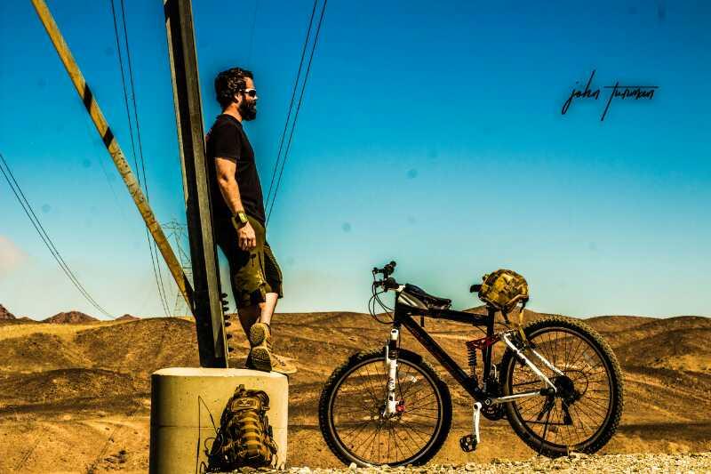 A person with a beard wearing a black shirt and shorts stands casually next to a power pole in a desert landscape, leaning against the pole. Beside them is a mountain bike with a camouflage backpack attached, set against a backdrop of dry, sandy hills and a clear blue sky. Sugarloaf Peak mountain bike trail.