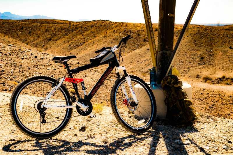 A black and white mountain bike is parked beside a rocky terrain under a tall pole. In the background, there are rolling hills and mountains, with a clear blue sky above. A backpack is resting against the pole, indicating a potential stop for a break during a biking adventure. Sugarloaf Peak mountain bike trail.