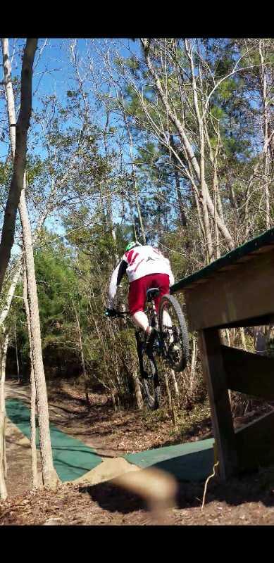 A person in a white and red outfit is performing a jump on a mountain bike over a wooden ramp, surrounded by trees. The ground beneath the bike is a green pathway, indicating a bike trail or park. The scene captures a moment of action and excitement in a natural setting. Horry County Bike Run Park mountain bike trail.
