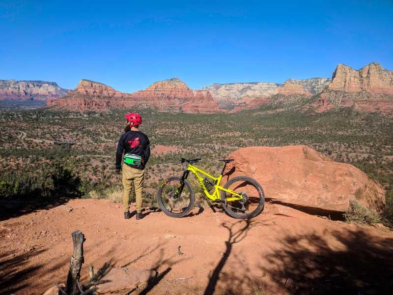A person in a red helmet and black jacket stands on a rocky ledge, looking out over a expansive desert landscape with red rock formations and blue sky. A bright yellow mountain bike is parked beside them. Hiline mountain bike trail.