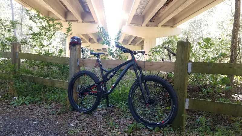 A mountain bike leaning against a wooden fence in a lush green area, with an overpass visible in the background. Sunlight filters through the structure, creating a warm, inviting atmosphere. A helmet is placed on the ground nearby. Fern Trail mountain bike trail.