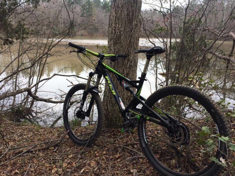 A black and green mountain bike is parked against a tree by a calm, muddy river. Surrounding the bike, there are bare trees and underbrush, indicative of a natural, wooded area. The scene is tranquil with a hint of winter fading into spring, suggesting an outdoor adventure environment. Itusi @ Lake Norman State Park mountain bike trail.