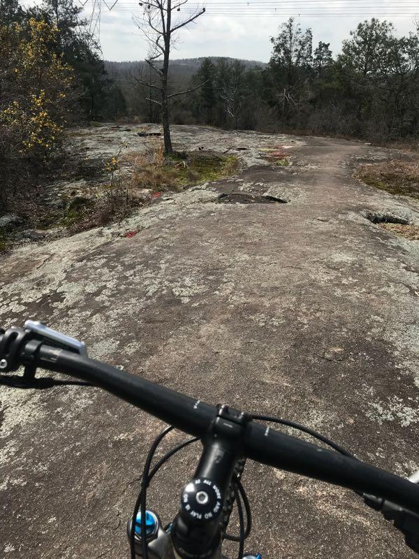 Mountain bike handlebars in the foreground with a rocky trail ahead, surrounded by sparse trees and a cloudy sky. The landscape includes patches of moss and scattered vegetation on the rocky surface. Clinton Nature Preserve mountain bike trail.