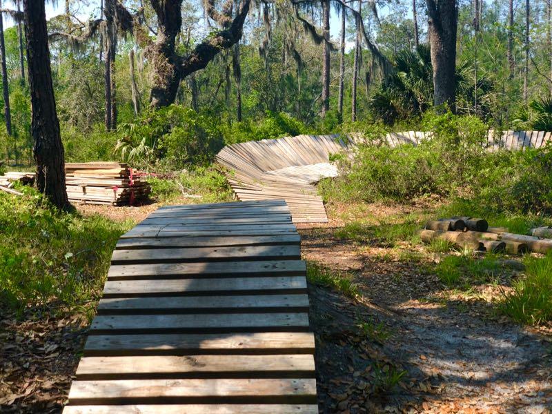 A winding wooden boardwalk through a forested area, surrounded by lush greenery and tall trees, with stacks of lumber visible on the left side of the path. Nocatee mountain bike trail.