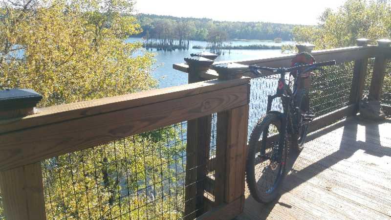 A mountain bike leaned against a wooden railing overlooking a scenic body of water surrounded by trees in early autumn. Tom Brown / Lafayette Heritage Park mountain bike trail.