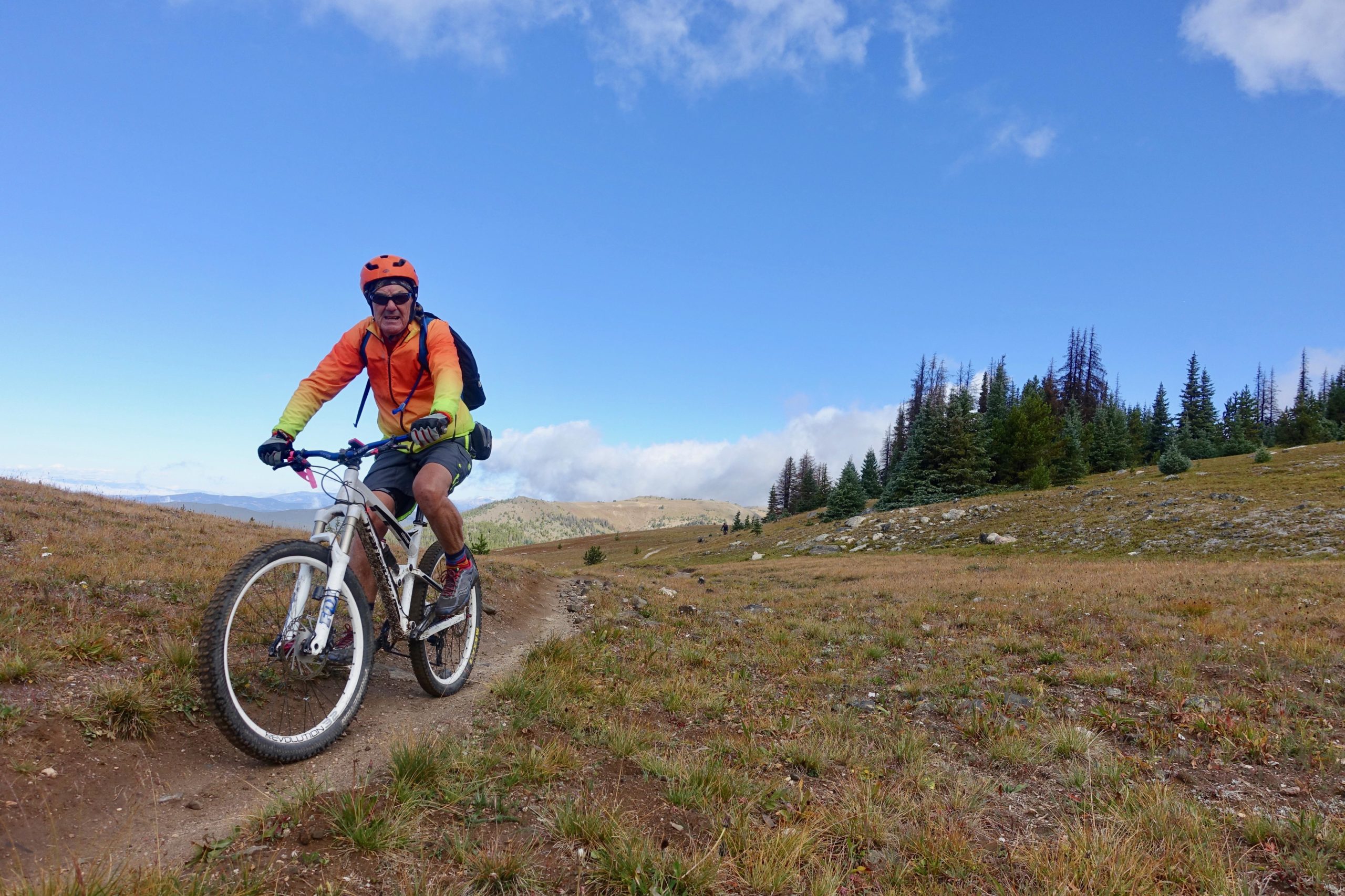 A middle-aged man riding a mountain bike on a grassy trail surrounded by rolling hills and a clear blue sky. He is wearing an orange and yellow long-sleeve shirt, a helmet, and shorts, and has a backpack on. The landscape features patches of trees in the background and a serene outdoor setting. Monarch Crest Trail mountain bike trail.