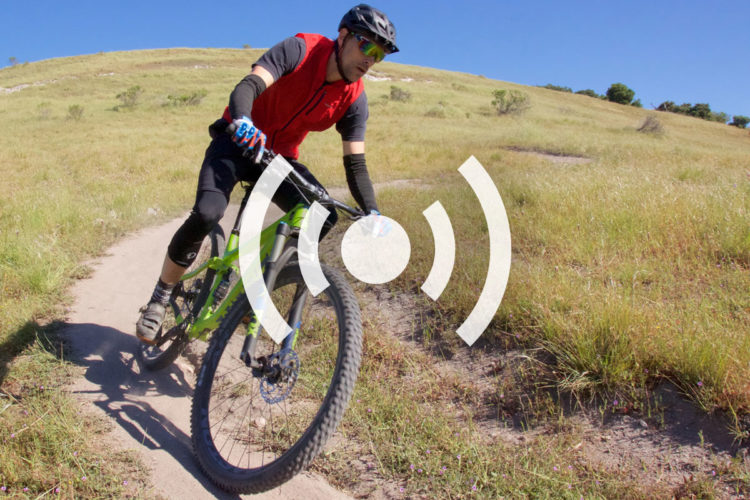 A person riding a green mountain bike on a dirt trail surrounded by grassy fields under a clear blue sky. The rider is wearing a red vest, black shorts, gloves, and sunglasses, focused on navigating the terrain.