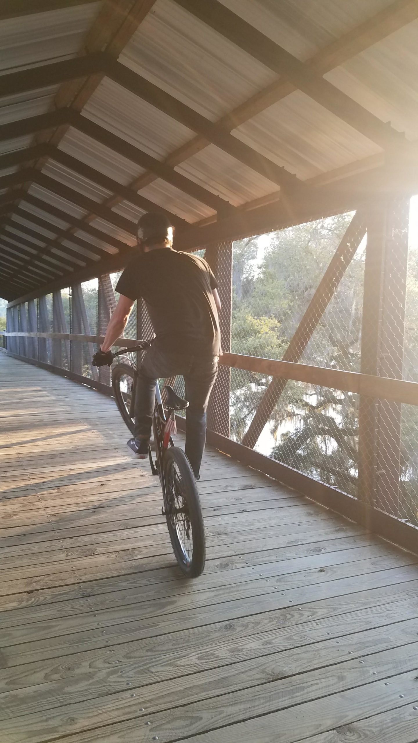 A person riding a bicycle on a wooden bridge, performing a wheelie. The scene is illuminated by soft sunlight, with trees visible in the background through the mesh fencing of the bridge. Tom Brown / Lafayette Heritage Park mountain bike trail.