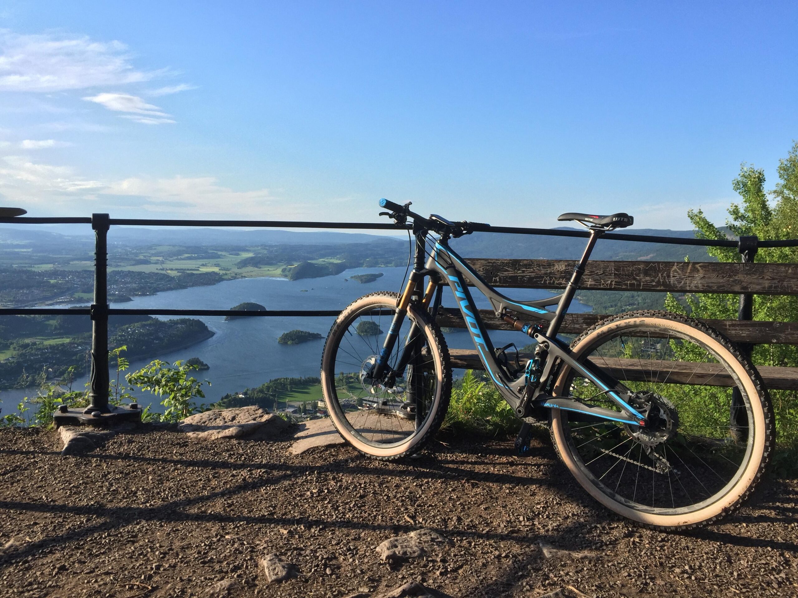 Pivot Mach 429SL Carbon: A mountain bike leaning against a wooden railing, overlooking a scenic view of a lake surrounded by greenery and hills under a clear blue sky.