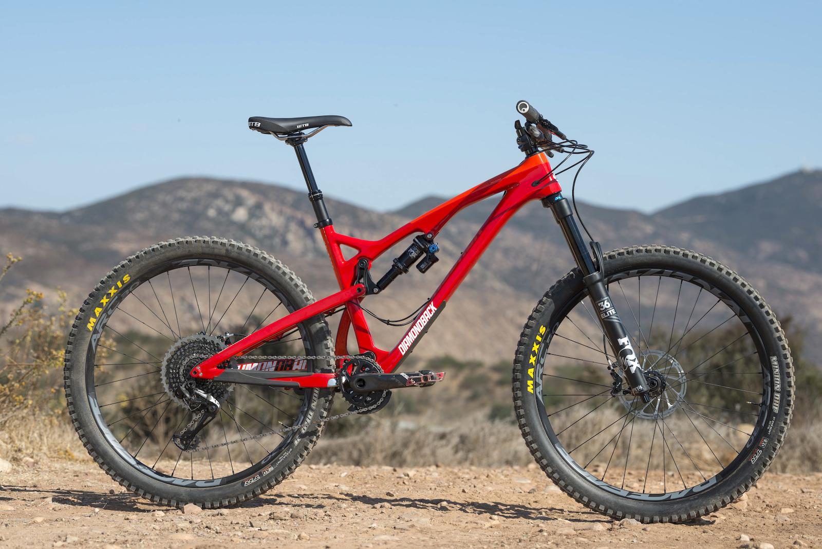 Diamondback Release 5c: A bright red mountain bike is positioned on a rocky outdoor trail, showcasing its sturdy frame and thick tires. The background features hills and a clear blue sky, emphasizing an adventurous and natural setting.