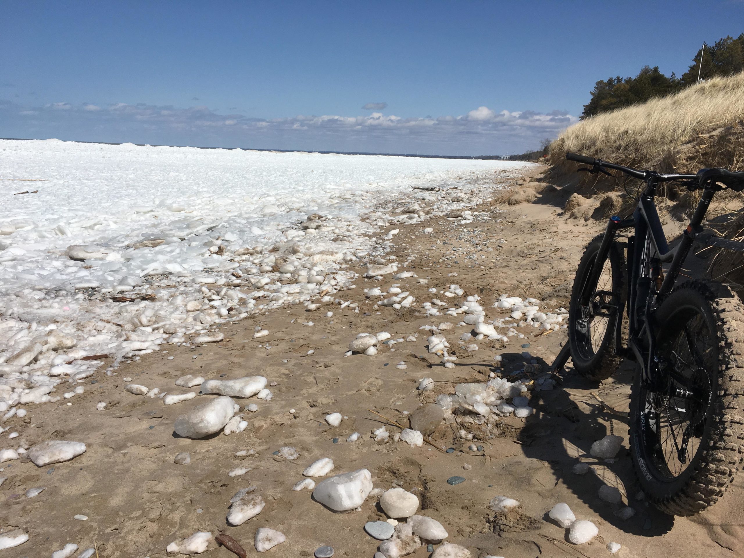 A sandy beach with scattered pieces of ice and foam along the shoreline, featuring a fat tire bicycle parked on the sand. In the background, there is a view of an icy lake under a clear blue sky with a few clouds. Dune grass lines the edge of the beach. Pinery Provincial Park mountain bike trail.