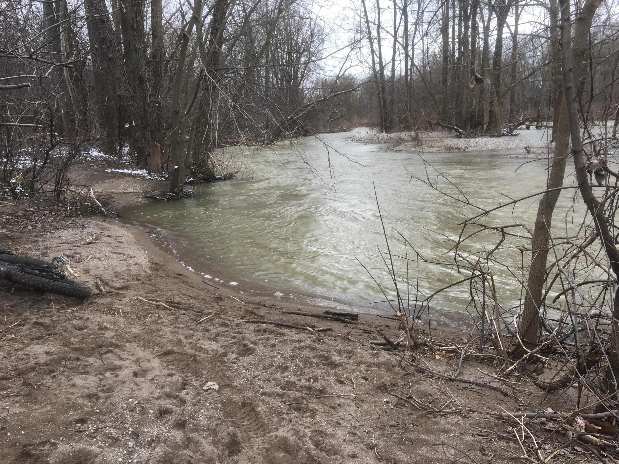 A riverside scene featuring a flowing river surrounded by bare trees and sparse vegetation. The sandy bank leads to the water's edge, which appears slightly muddy. In the foreground, there are traces in the sand and a discarded tire partially submerged in the sand. The overall atmosphere is tranquil and slightly overcast, hinting at early spring or late winter conditions. Western University trails mountain bike trail.