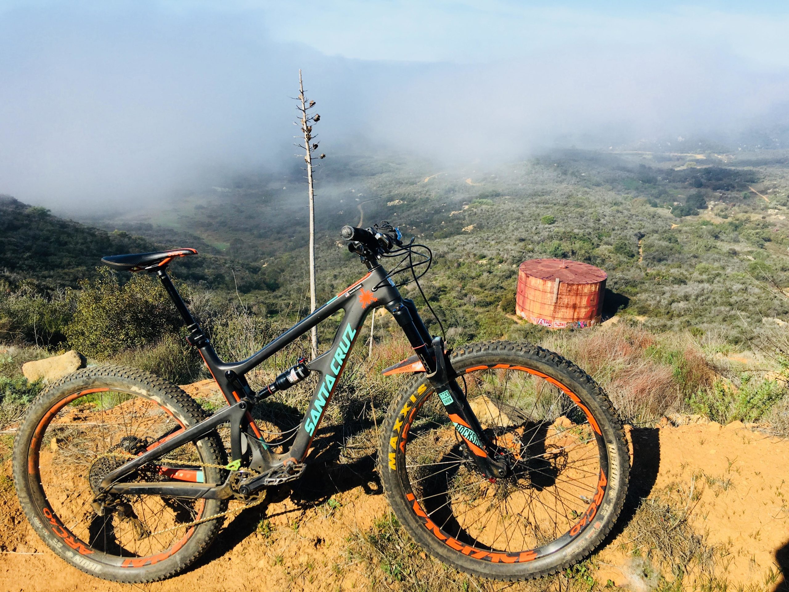 A mountain bike with a black frame and orange accents is positioned on a dirt trail overlooking a misty landscape. In the background, rolling hills are partially obscured by fog, and a large, rusted water tank can be seen in the distance. The scene captures the beauty of nature and the thrill of outdoor biking. Daley Ranch mountain bike trail.