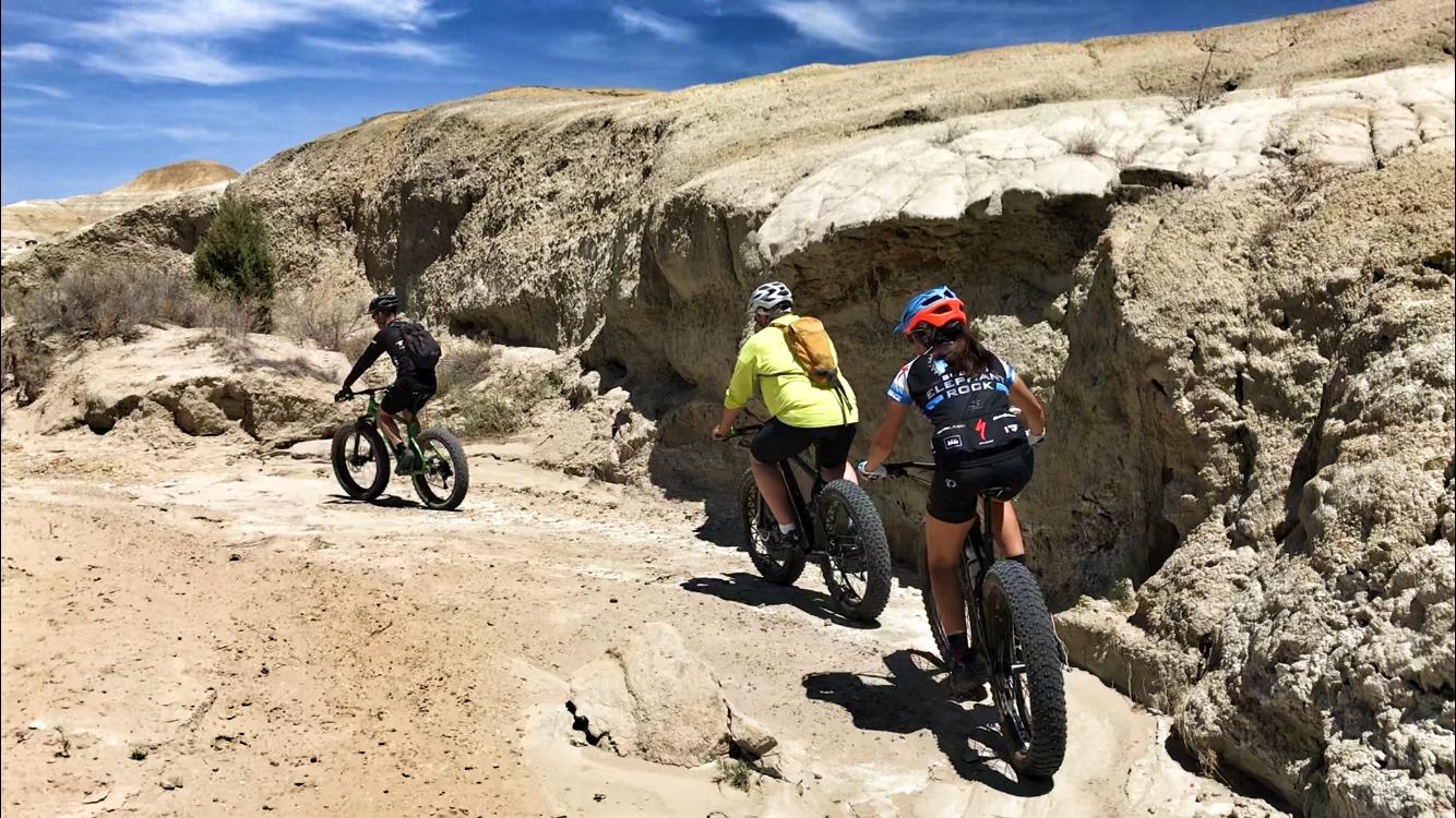 Three individuals riding fat bikes along a sandy trail in a rocky landscape under a blue sky. The terrain features light-colored hills and rugged cliffs, with shrubs scattered around. The cyclists are wearing helmets and appropriate biking gear. Double Slot Fat Bike Trail mountain bike trail.