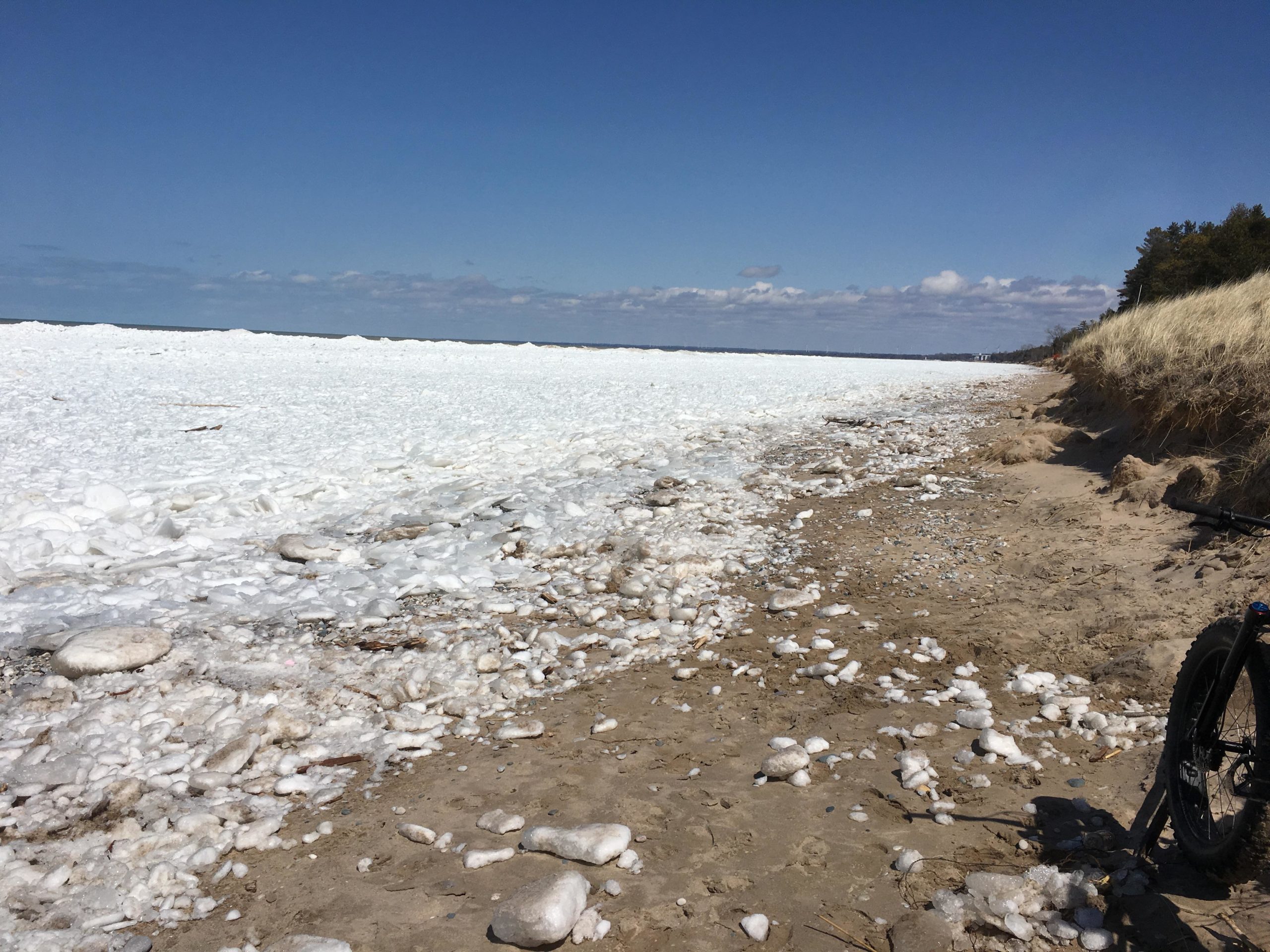 A shoreline scene featuring a sandy beach with scattered white foam and ice along the water's edge. In the background, a calm lake with a cloudy blue sky above and a strip of forested land visible on the right side. A bicycle is partially visible in the foreground. Pinery Provincial Park mountain bike trail.
