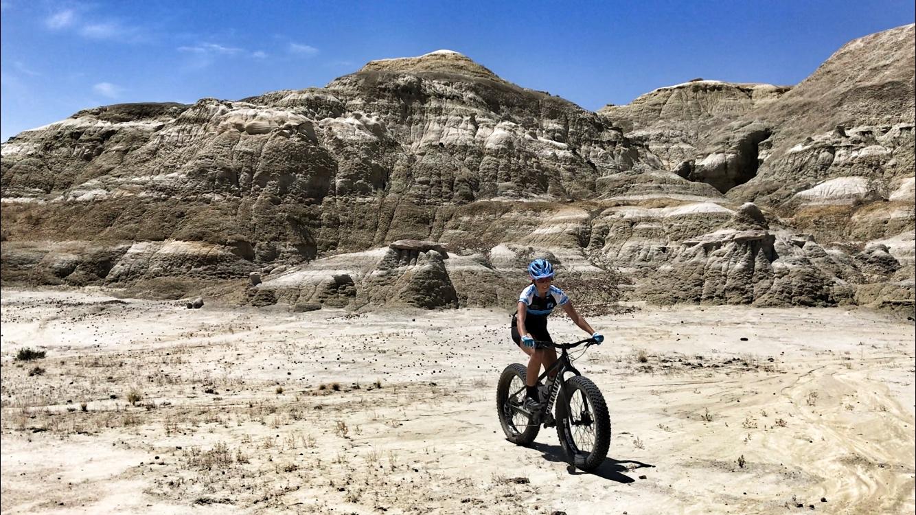 A cyclist riding a fat tire bike across a dry, rocky landscape with unique, layered geological formations under a clear blue sky. Double Slot Fat Bike Trail mountain bike trail.