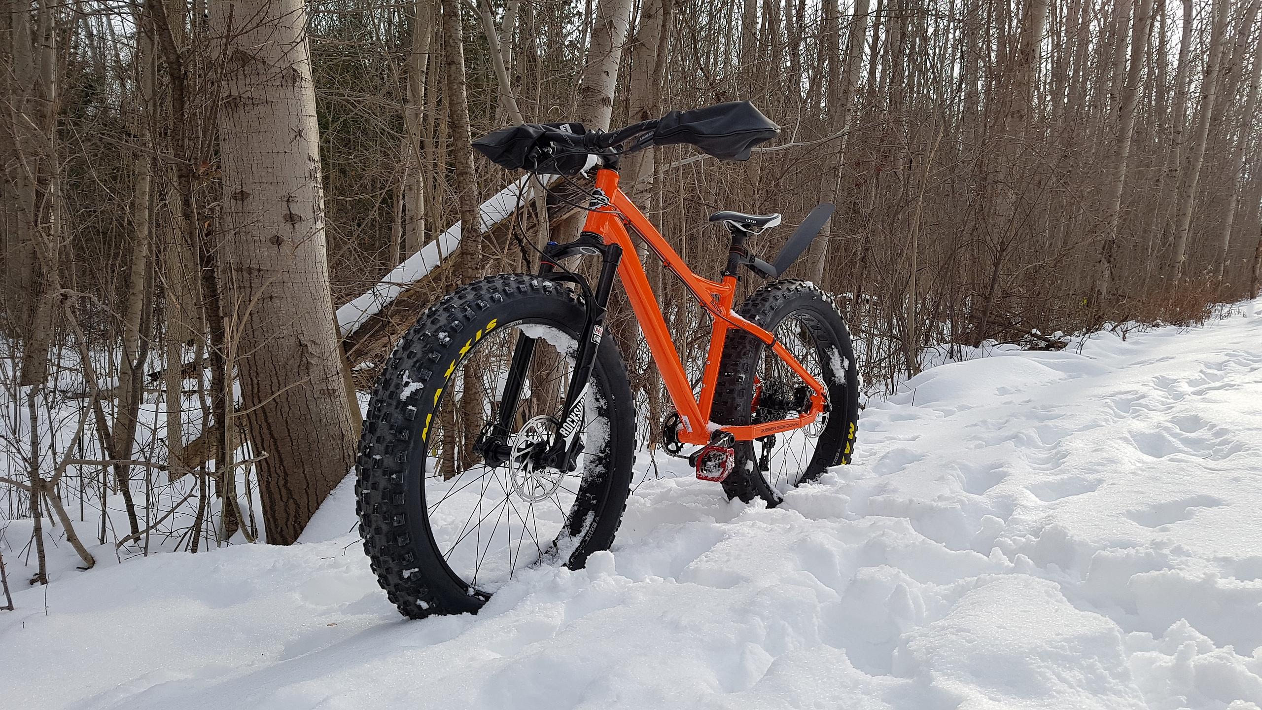RSD Bikes Mayor Aluminum: A bright orange fat bike resting on snow in a wooded area, with tall trees in the background and a trail visible in the snow. The bike features wide tires designed for snowy conditions.