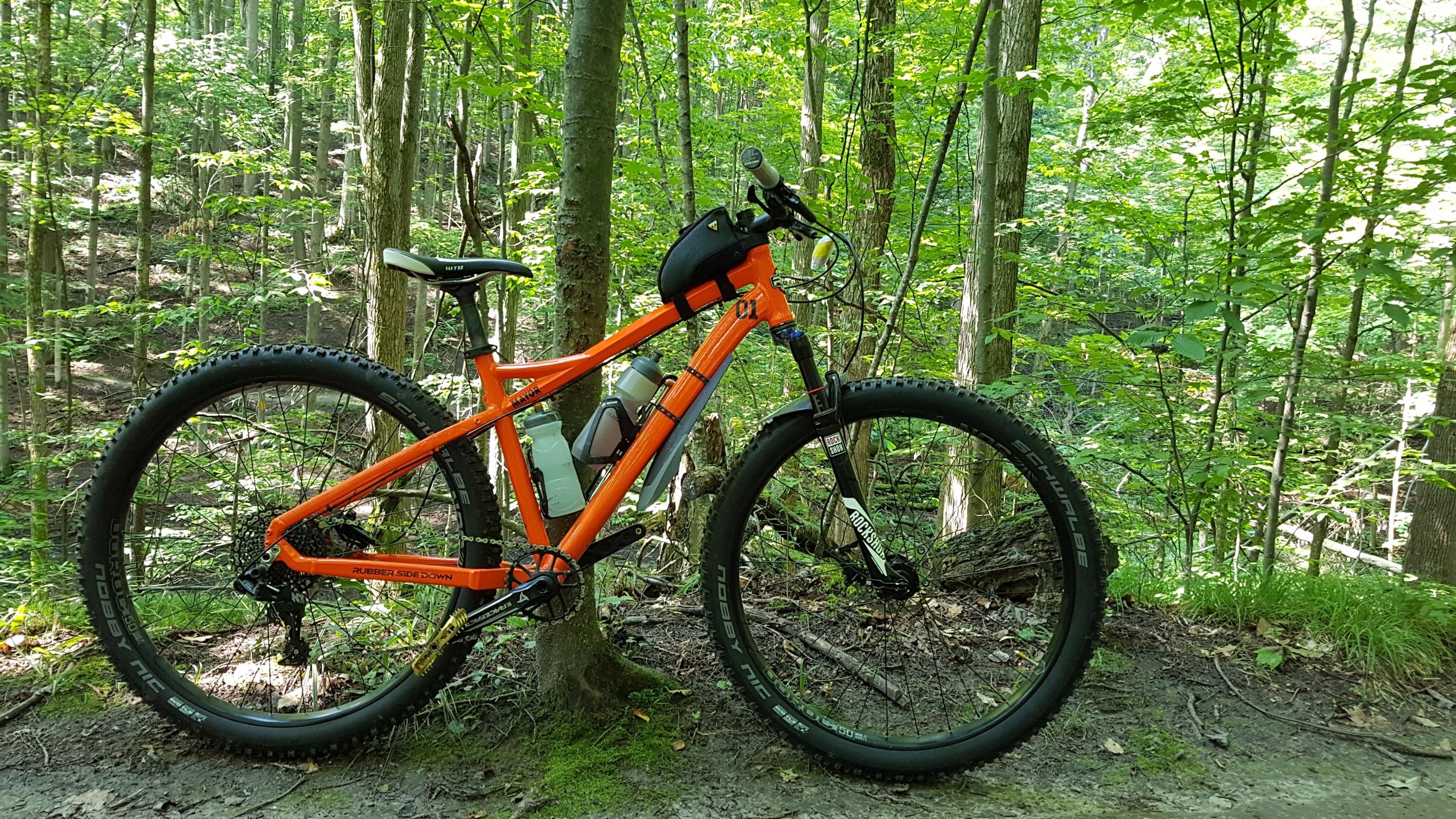 RSD Bikes Mayor Aluminum: A bright orange mountain bike resting on a dirt path surrounded by lush green trees and foliage in a forested area. The bike features wide tires, a water bottle holder, and a small bag on the handlebars. Soft sunlight filters through the leaves, creating a serene outdoor atmosphere.