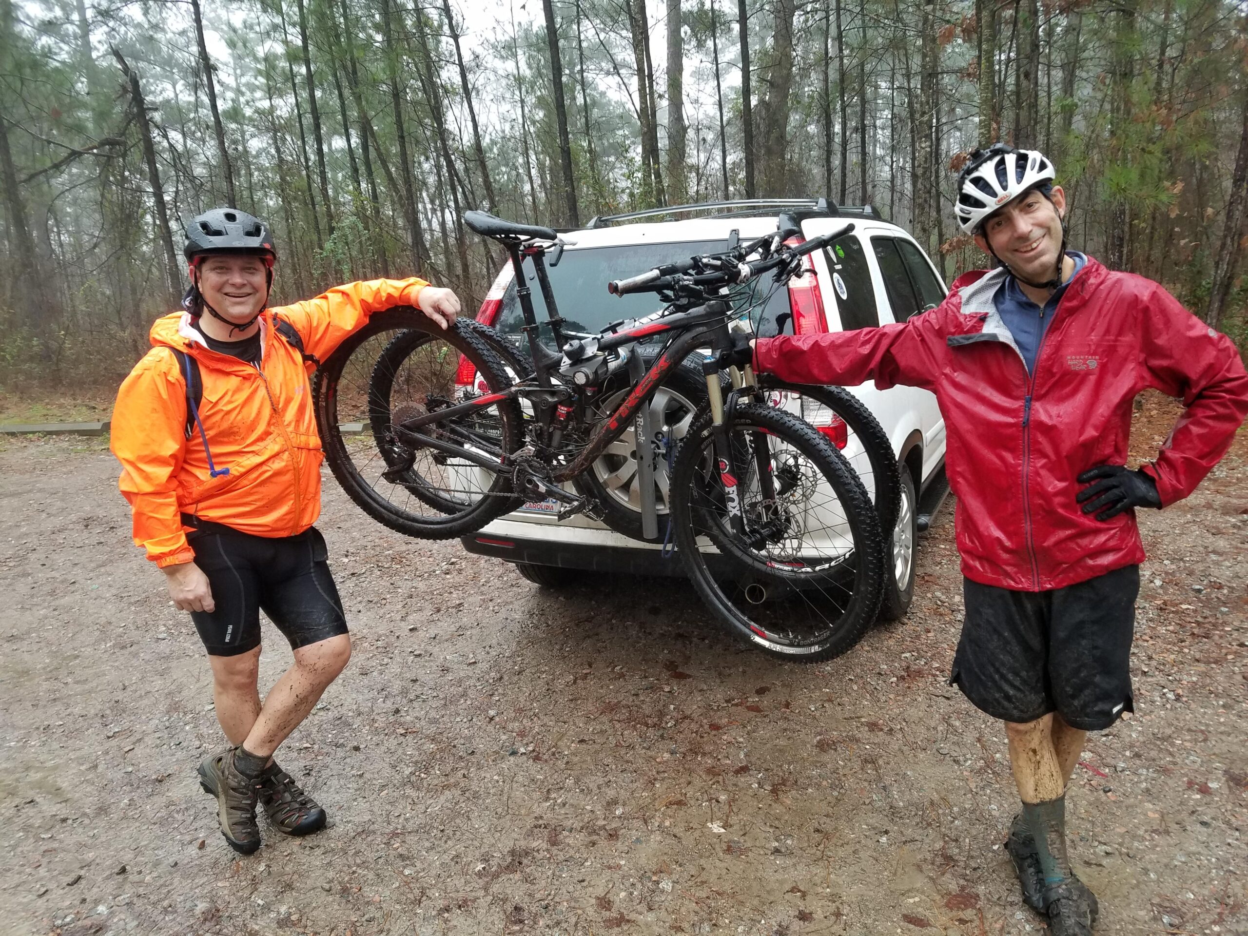 Trek Fuel EX 8: Two mountain bikers standing beside an SUV in a forested area. One man is wearing a bright orange waterproof jacket and black shorts, holding a bike attached to a rack on the vehicle. The other man is in a red waterproof jacket and black shorts, smiling while resting his hand on the bike. Both appear muddy, indicating they have been biking in wet conditions. The scene is set in a rainy, wooded environment.