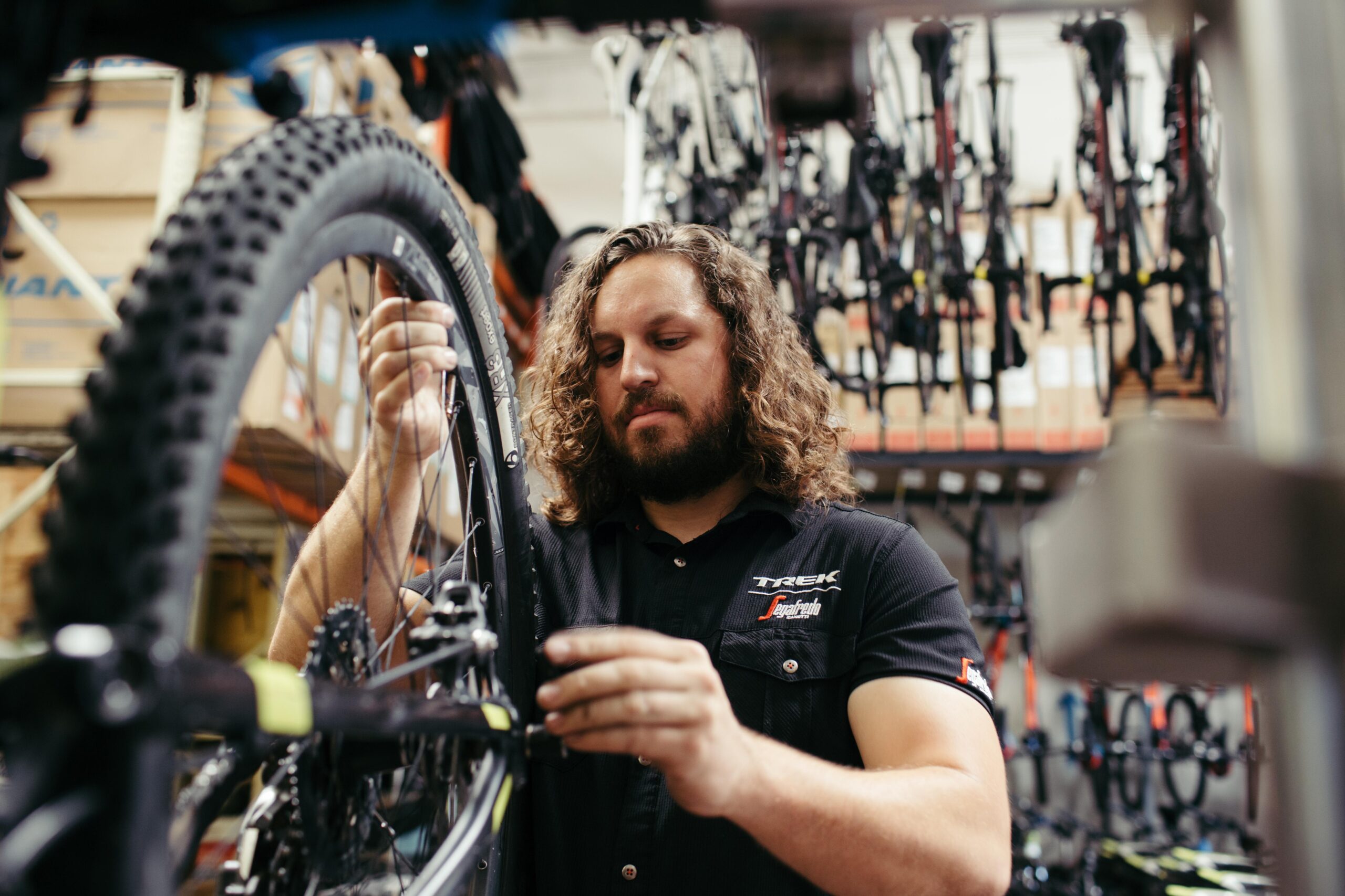 A mechanic with curly hair works on a bicycle in a workshop filled with bike parts and hanging bicycles in the background. The mechanic is focused on adjusting the bike's wheel, wearing a black shirt with the Trek logo.