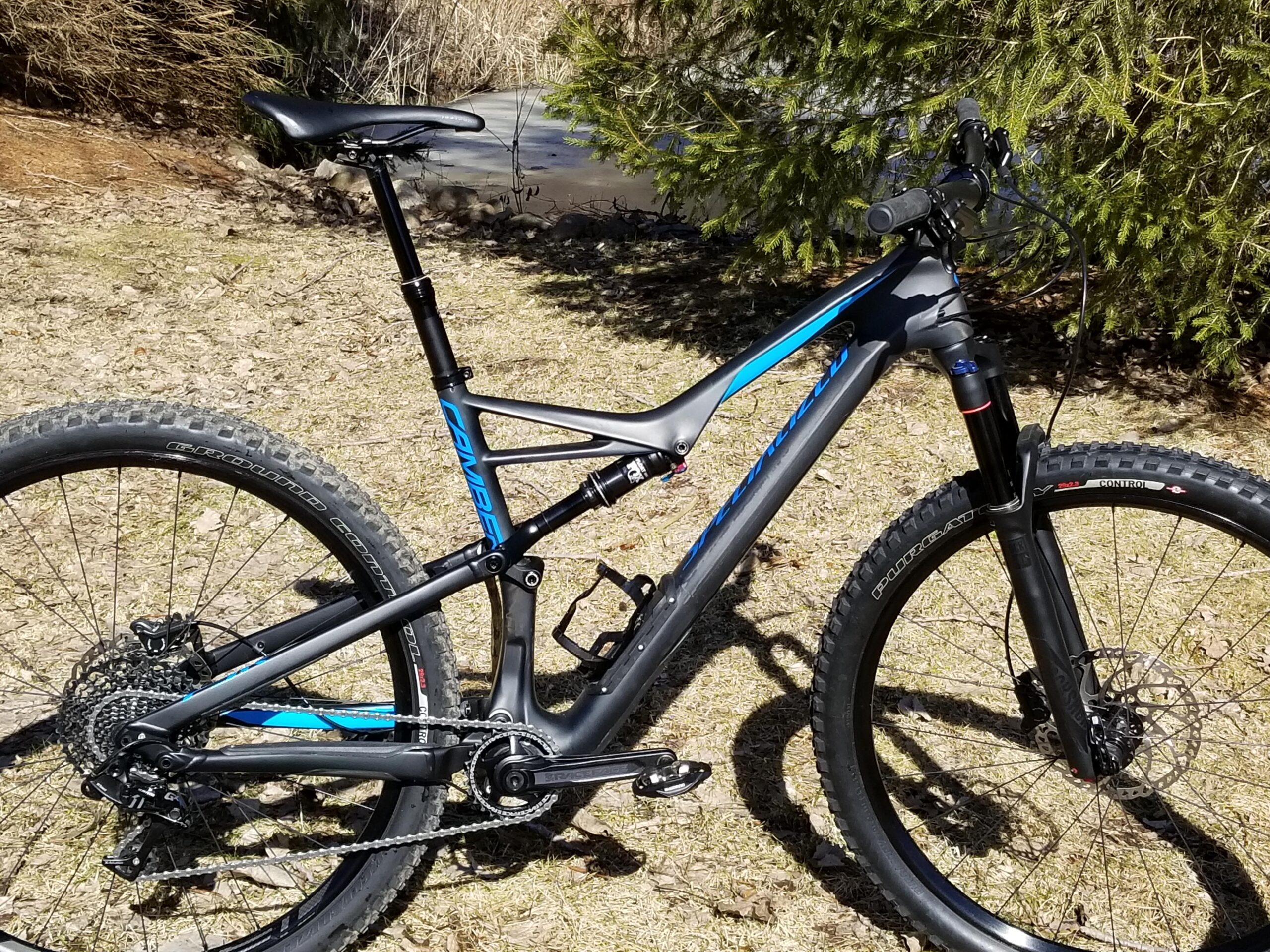 Specialized Camber Comp 29: A mountain bike with a sleek black and blue design is positioned on grass, featuring thick tires and a prominent suspension system. The background includes trees and a hint of water, indicating an outdoor setting.