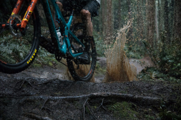 A close-up shot of a mountain bike maneuvering through a muddy trail in a forest, with water splashing up from the rear wheel. The bike features bright orange and teal colors, and the background showcases lush greenery and tall trees, highlighting the rugged outdoor environment.