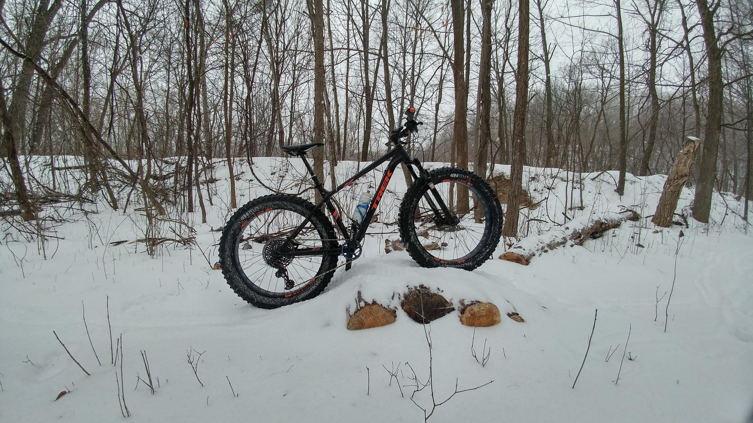 A black fat bike with orange accents is positioned on a small mound surrounded by snow-covered ground and bare trees. The scene is serene, with a gray sky above, suggesting winter conditions in a forested area. Glacial Blue Hills mountain bike trail.