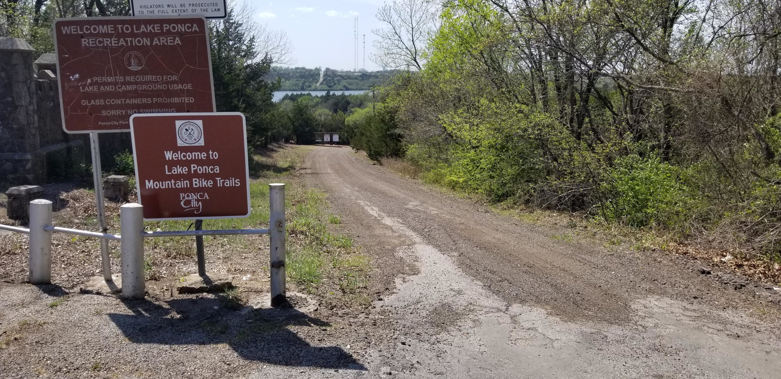 A dirt road leading into a wooded area, with two signs at the entrance. One sign indicates the welcome to Lake Ponca Recreation Area, detailing regulations like permit requirements and prohibitions on glass containers. The second sign welcomes visitors to Lake Ponca Mountain Bike Trails, marked with the Ponca City logo. In the background, a glimpse of the lake can be seen through the trees. Lake Ponca Trails mountain bike trail.