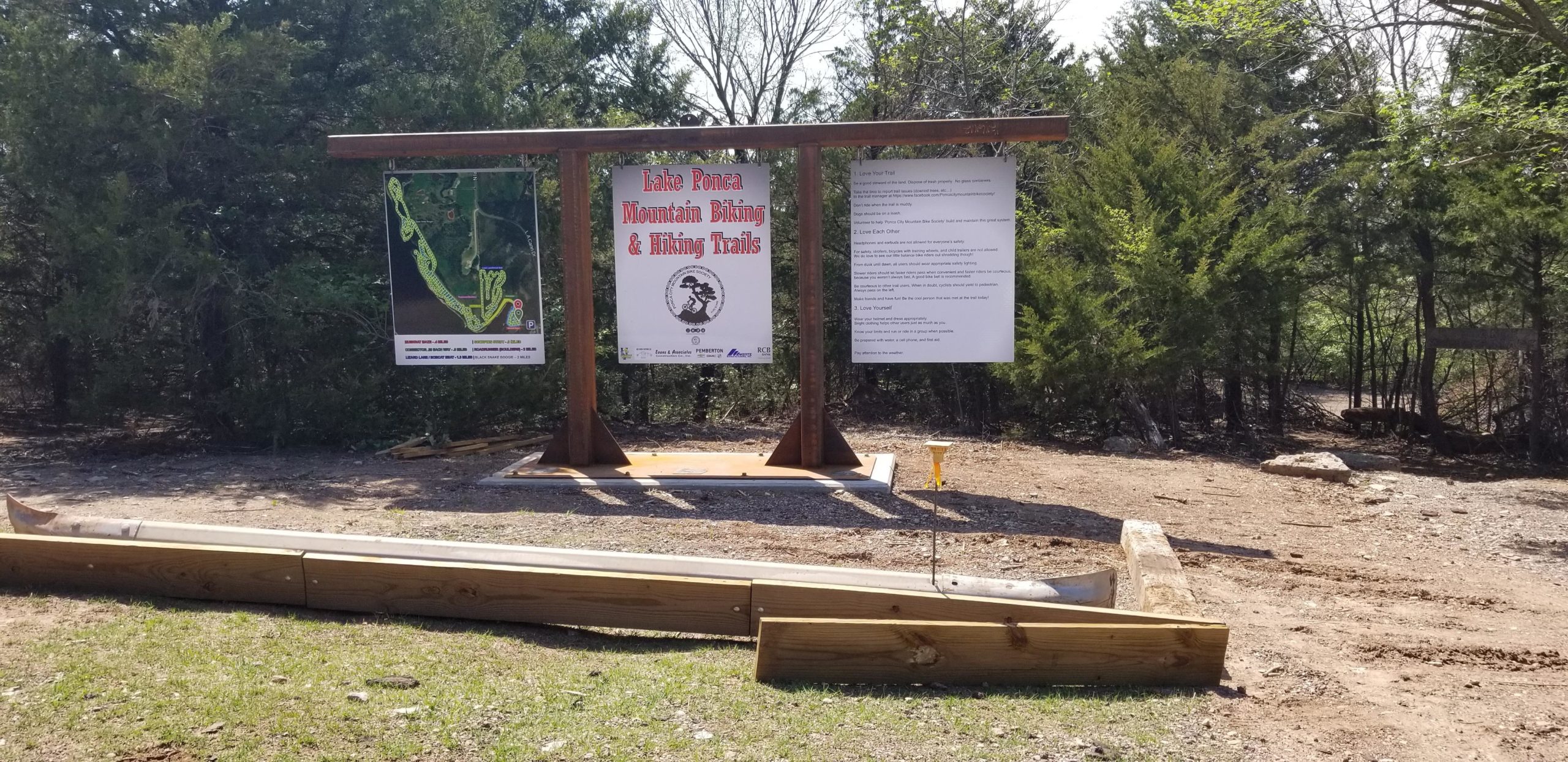 Signpost displaying information about Lake Ponca Mountain Biking and Hiking Trails, including a map and guidelines for trail users. Surrounding the sign are trees and natural landscape, with some construction materials visible in the foreground. Lake Ponca Trails mountain bike trail.