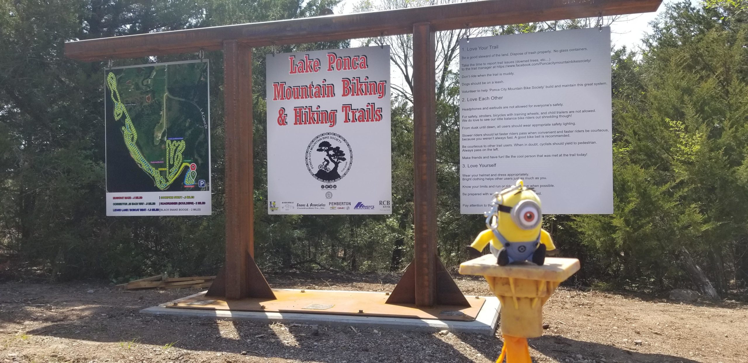 A signpost at Lake Ponca displaying a trail map for mountain biking and hiking, with detailed instructions and guidelines for trail users. A small toy figure resembling a Minion is perched on a wooden post in the foreground, while trees are visible in the background. Lake Ponca Trails mountain bike trail.