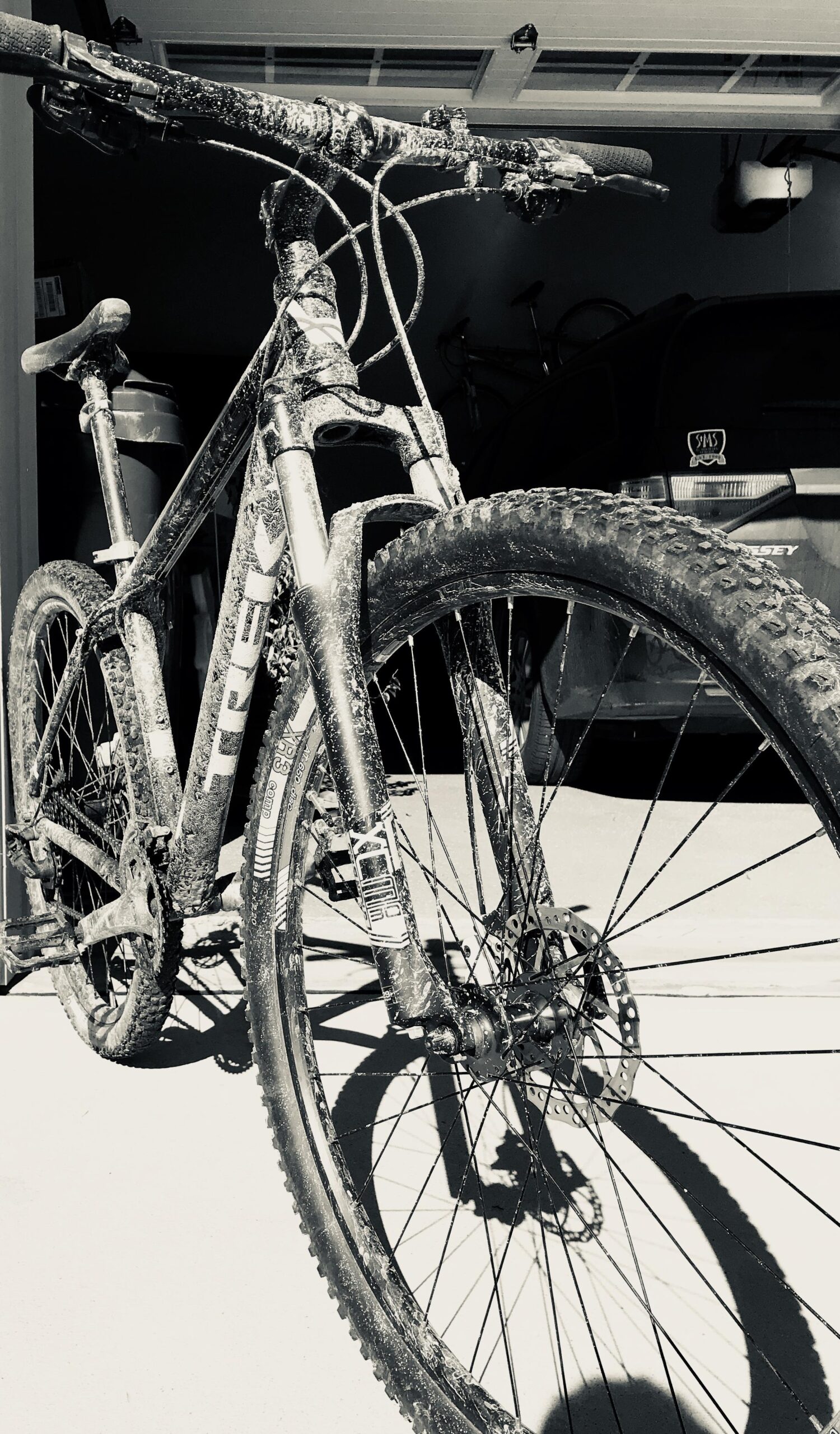 Trek Marlin 5 29er: A close-up image of a muddy mountain bike, showcasing its front wheel, frame, and handlebars. The bike is positioned in a garage setting, with a dark background that includes a glimpse of another bike and a vehicle. The monochrome effect highlights the dirt and grime accumulated on the bike from recent outdoor use.