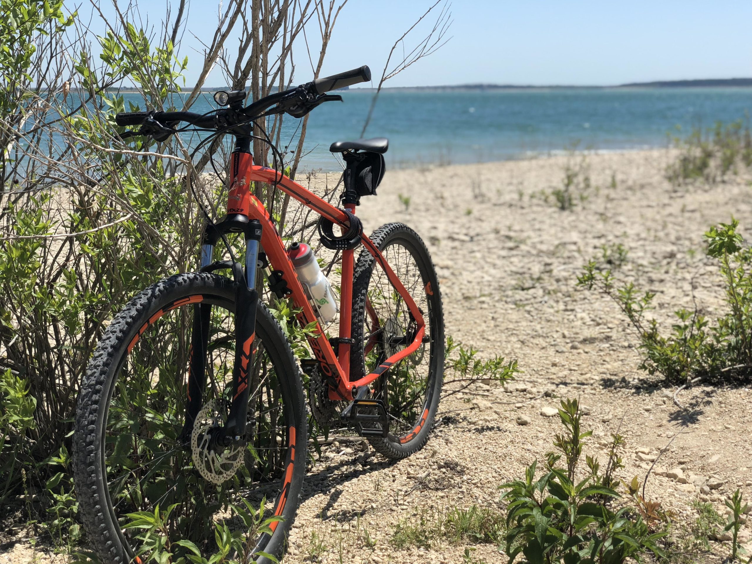 A bright orange mountain bike leaning against sparse vegetation near a sandy beach, with a clear blue body of water in the background and a bright sky. Dana Peak mountain bike trail.