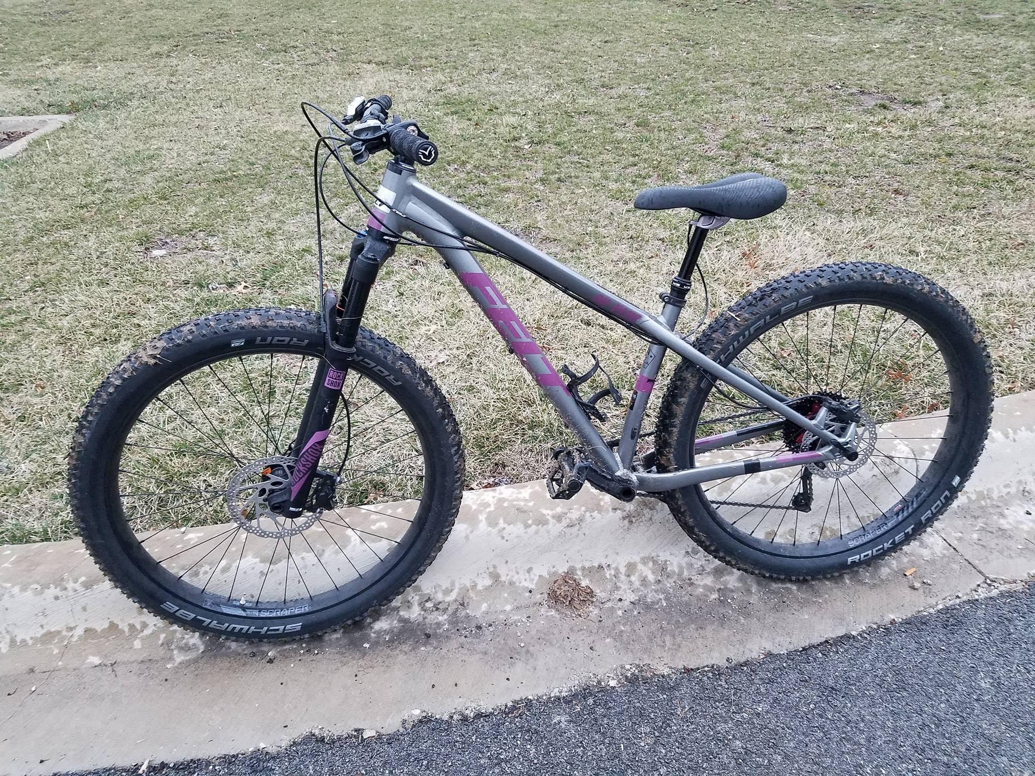 Felt Surplus: A gray mountain bike with purple accents, resting on a concrete curb near a grassy area. The bike features wide tires with a tread pattern suitable for off-road terrain, and the frame has a sturdy design. The handlebars are equipped with a brake and gear shifters.