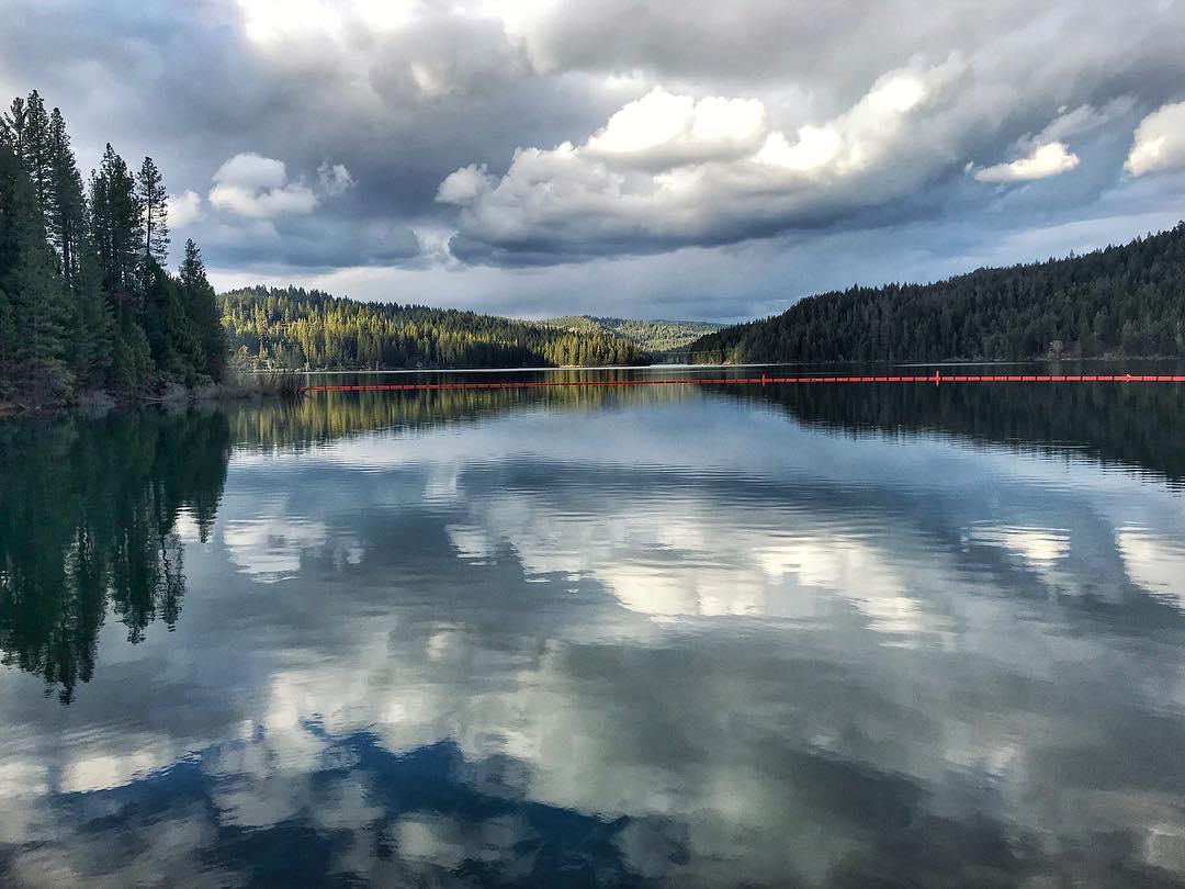 A serene landscape featuring a calm lake surrounded by lush green trees, with a cloudy sky reflected in the water. A bright red barrier is visible across the lake in the distance, enhancing the tranquil atmosphere of the scene. Sly Park mountain bike trail.