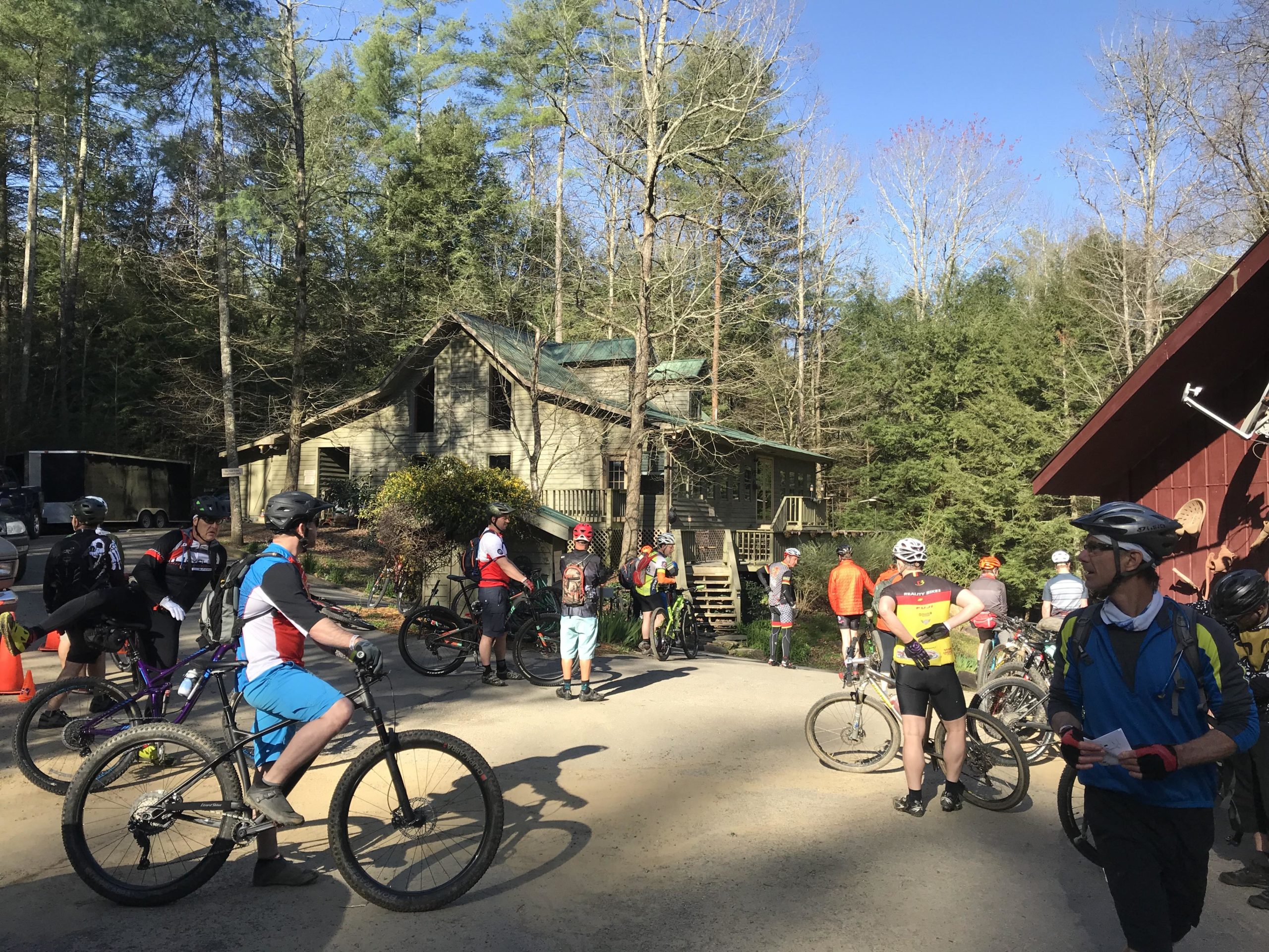 A group of mountain bikers gathers at a rustic outdoor location surrounded by trees. The scene shows riders in colorful cycling gear standing next to their bikes, with a wooden building in the background. It is a clear day with blue skies, suggesting a vibrant outdoor biking event. Pinhoti: Tatum Lead mountain bike trail.