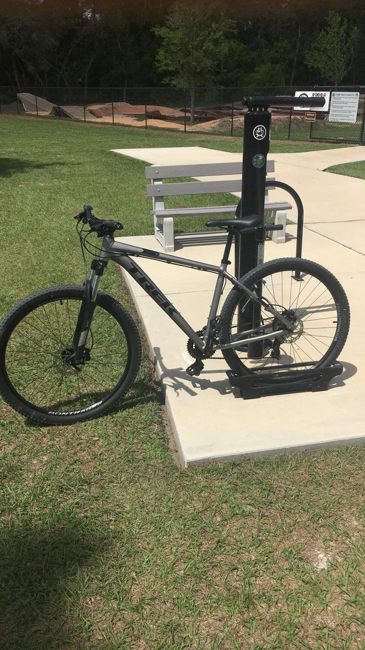 Trek Marlin 5: A black Trek mountain bike parked on a concrete platform near a grassy area, with a bike repair station beside it. In the background, there is a fenced area with dirt ramps for biking. A bench is visible on the platform, and trees are in the background.