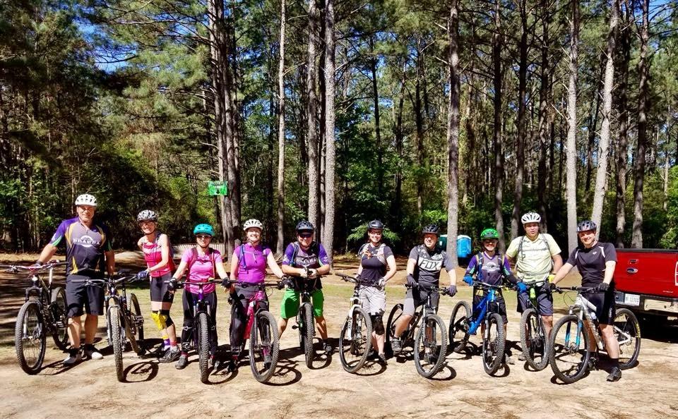 A group of ten diverse mountain bikers posing with their bikes in a wooded area, surrounded by tall pine trees. They are wearing helmets and various biking attire, and some bikes have distinctive colors. The atmosphere is lively and energetic, suggesting a day of outdoor adventure.
