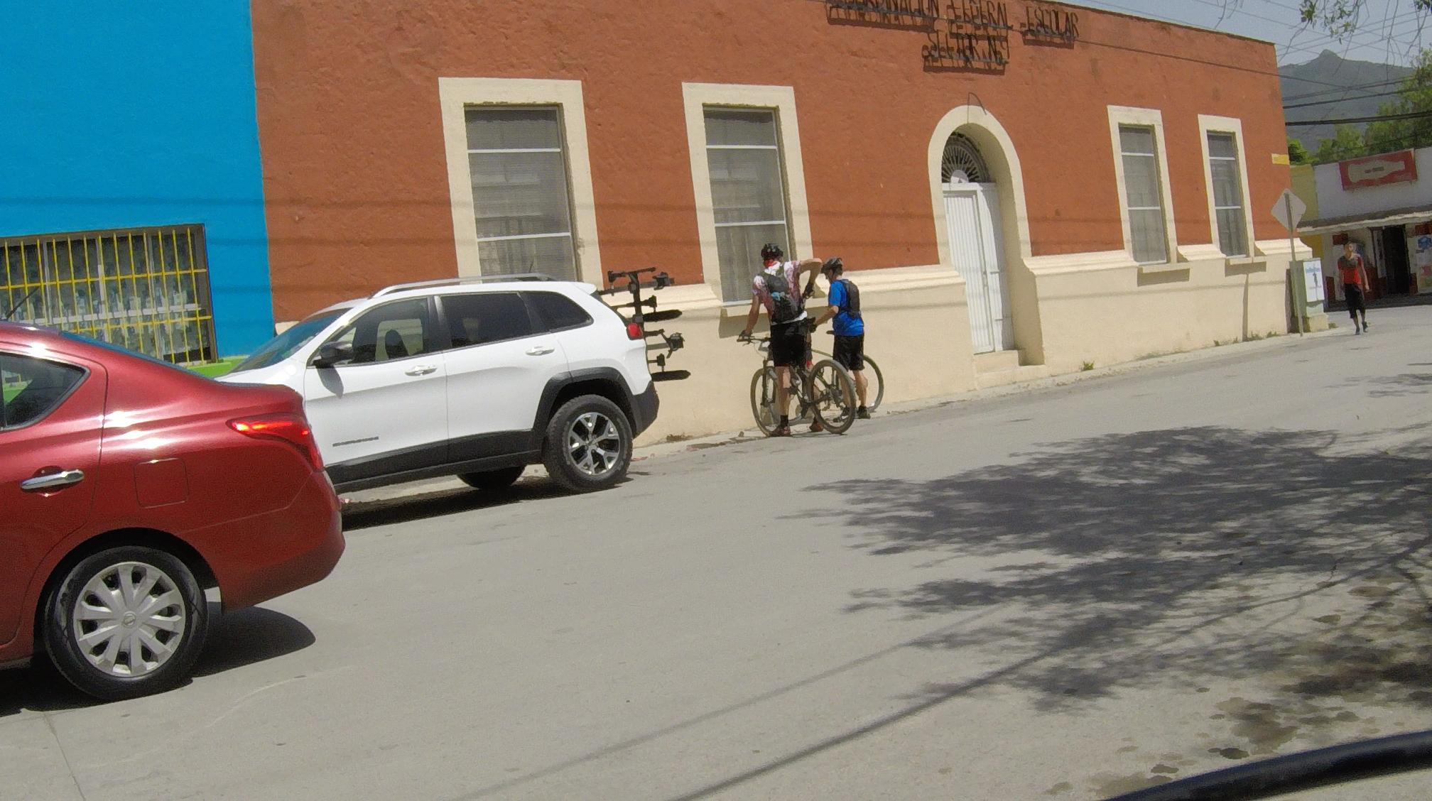 Two cyclists are conversing next to their bikes near a red and blue building. A white SUV with a bike rack is parked nearby, while a red car is in the foreground. A pedestrian is walking along the road in the background. Bosque Encantado mountain bike trail.