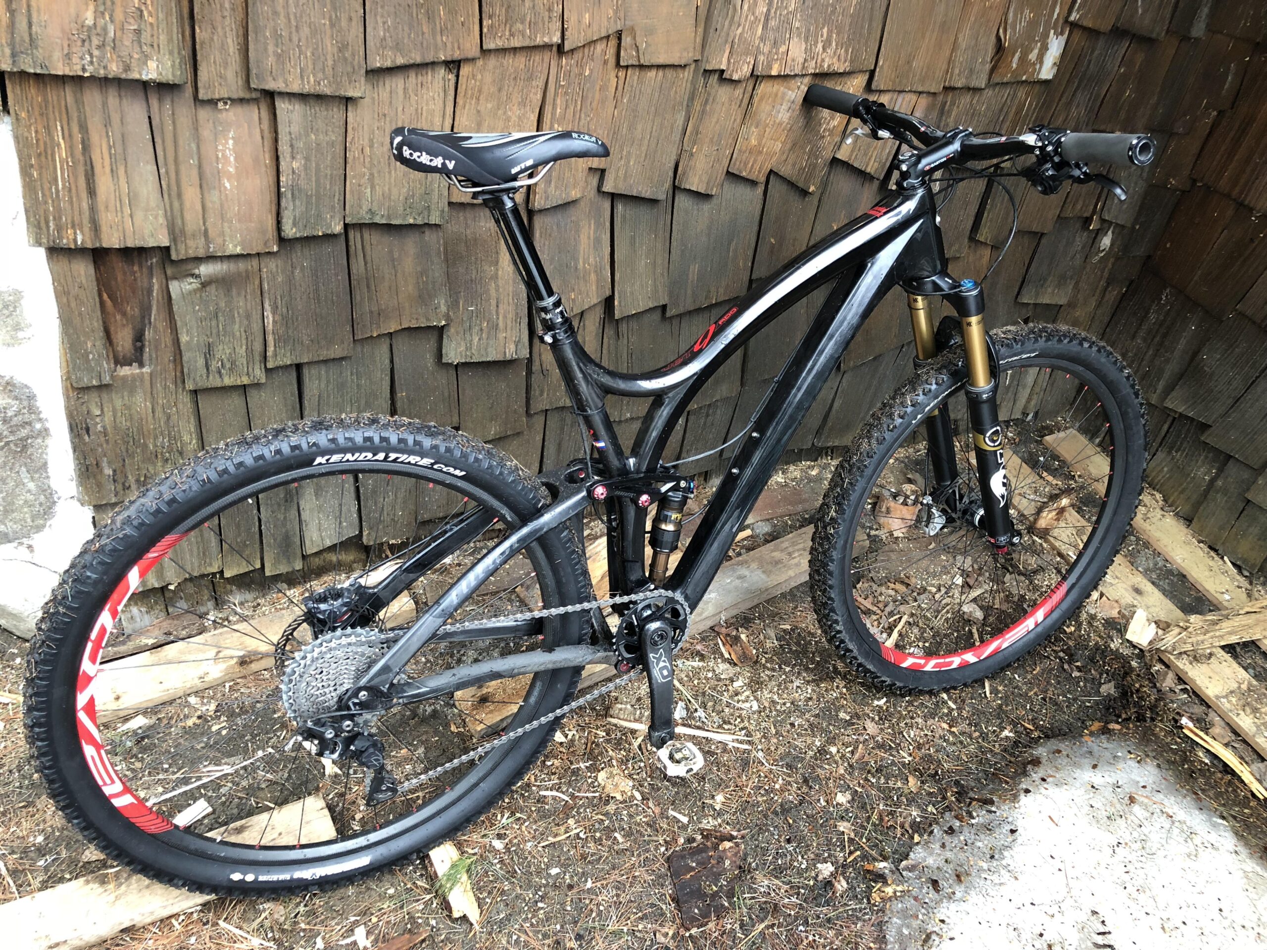 Niner Jet 9 RDO: A black mountain bike with red accents, leaning against a wooden wall. The bike features a dual suspension system, knobby tires, and a gear system with a cassette visible on the rear wheel. The ground is covered with dirt and wooden planks.