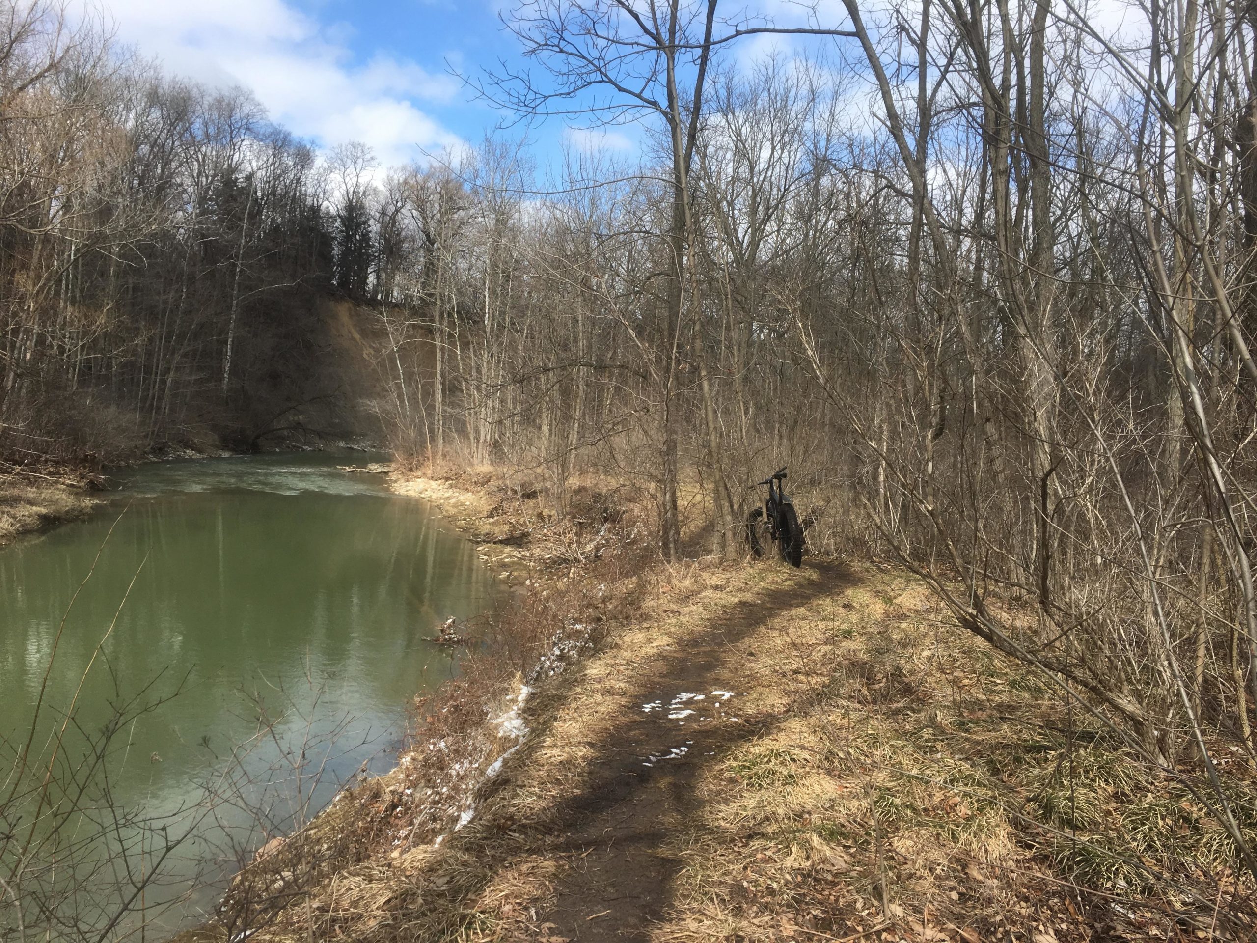 A peaceful outdoor scene depicting a winding dirt path alongside a calm, green river, bordered by bare trees. A bicycle is leaned against a tree on the right, and patches of snow and dry grass are visible on the ground, suggesting early spring conditions. The sky is partly cloudy, adding to the tranquil atmosphere of the landscape. Western University trails mountain bike trail.