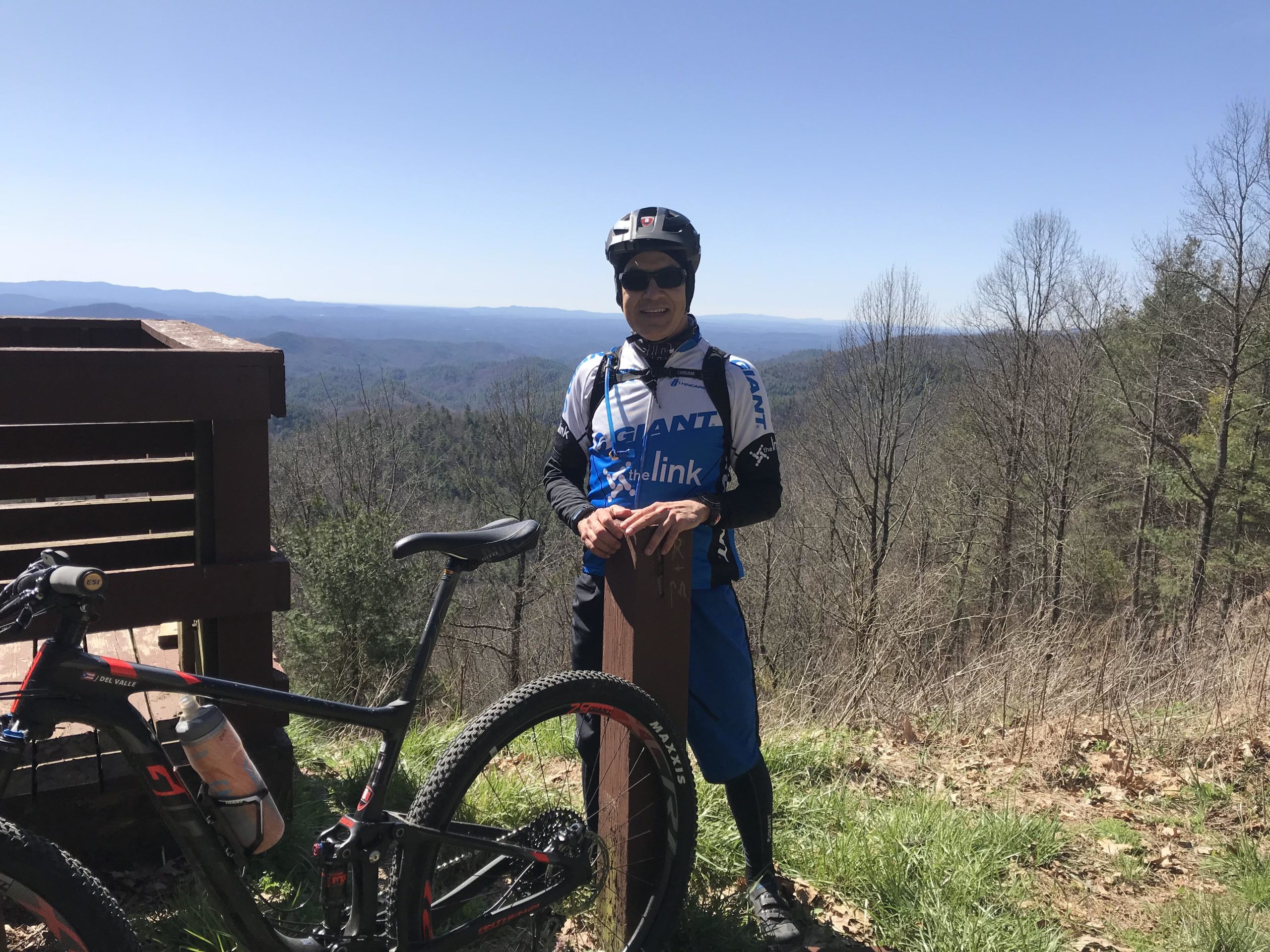 A person in a cycling outfit stands beside a mountain bike at a scenic overlook. The background features rolling hills and a clear blue sky. The cyclist is resting against a wooden post, enjoying the view of the landscape. Pinhoti: Tatum Lead mountain bike trail.