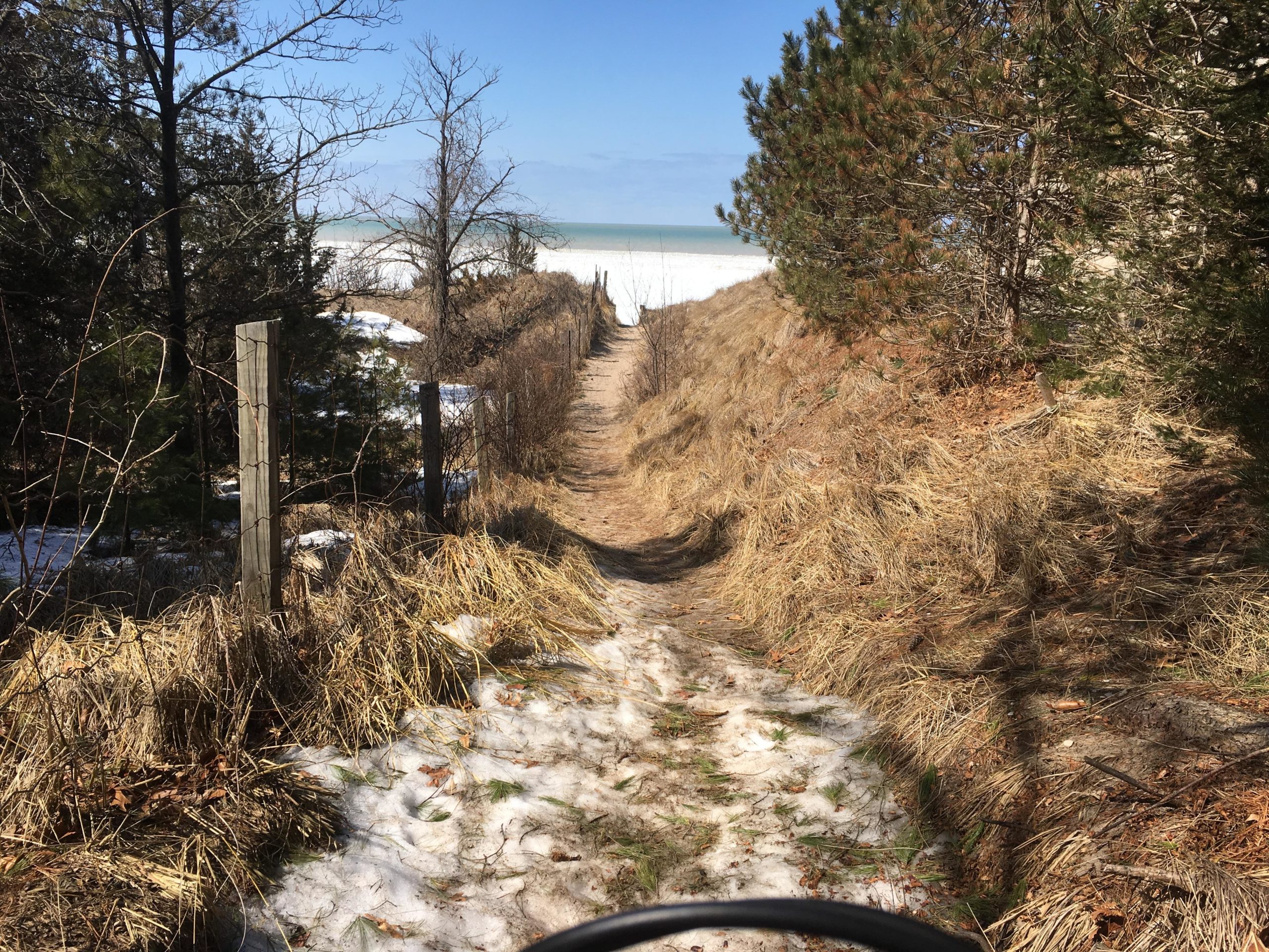 A sandy path leads through a mix of grass and, in some areas, remnants of snow, bordered by sparse trees and a wooden fence. The path stretches toward a bright, clear sky and the distant shoreline of a lake or ocean, hinting at a scenic view ahead. Pinery Provincial Park mountain bike trail.