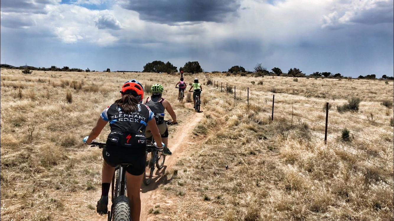 A group of mountain bikers riding along a dirt trail through a dry, grassy landscape under a partly cloudy sky. The scene shows five cyclists, some in colorful helmets and jerseys, biking away from the camera towards the horizon. A wire fence runs parallel to the trail on the right. The environment appears warm and sunny, with sparse vegetation. Alien Run Trail mountain bike trail.
