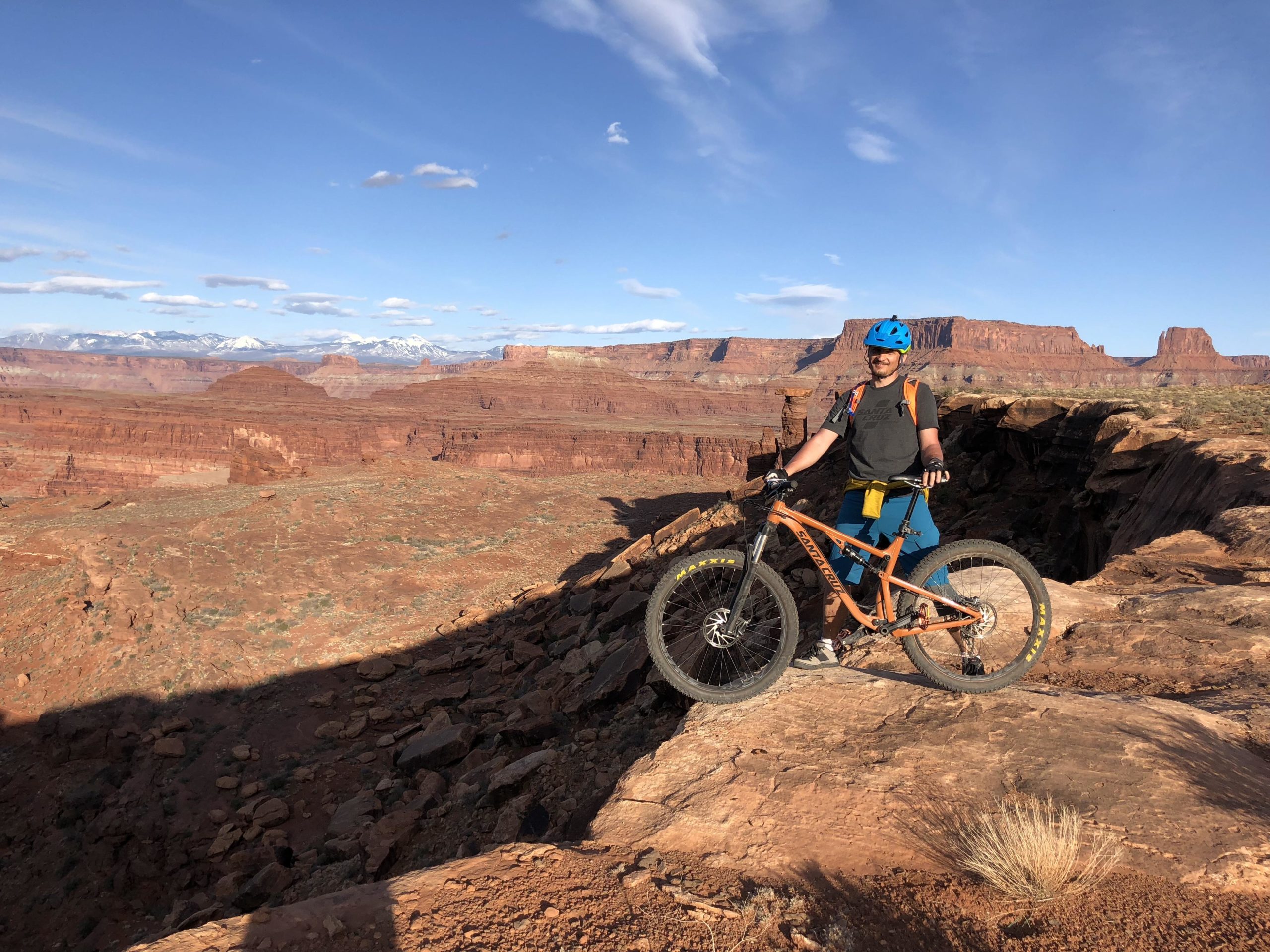 A person standing beside a mountain bike at the edge of a rocky cliff, overlooking a vast canyon landscape with red rock formations and snow-capped mountains in the background. The sky is clear with a few clouds. The individual is wearing a blue helmet, a gray shirt, and blue shorts, with a backpack and a jacket tied around their waist. White Rim Trail mountain bike trail.