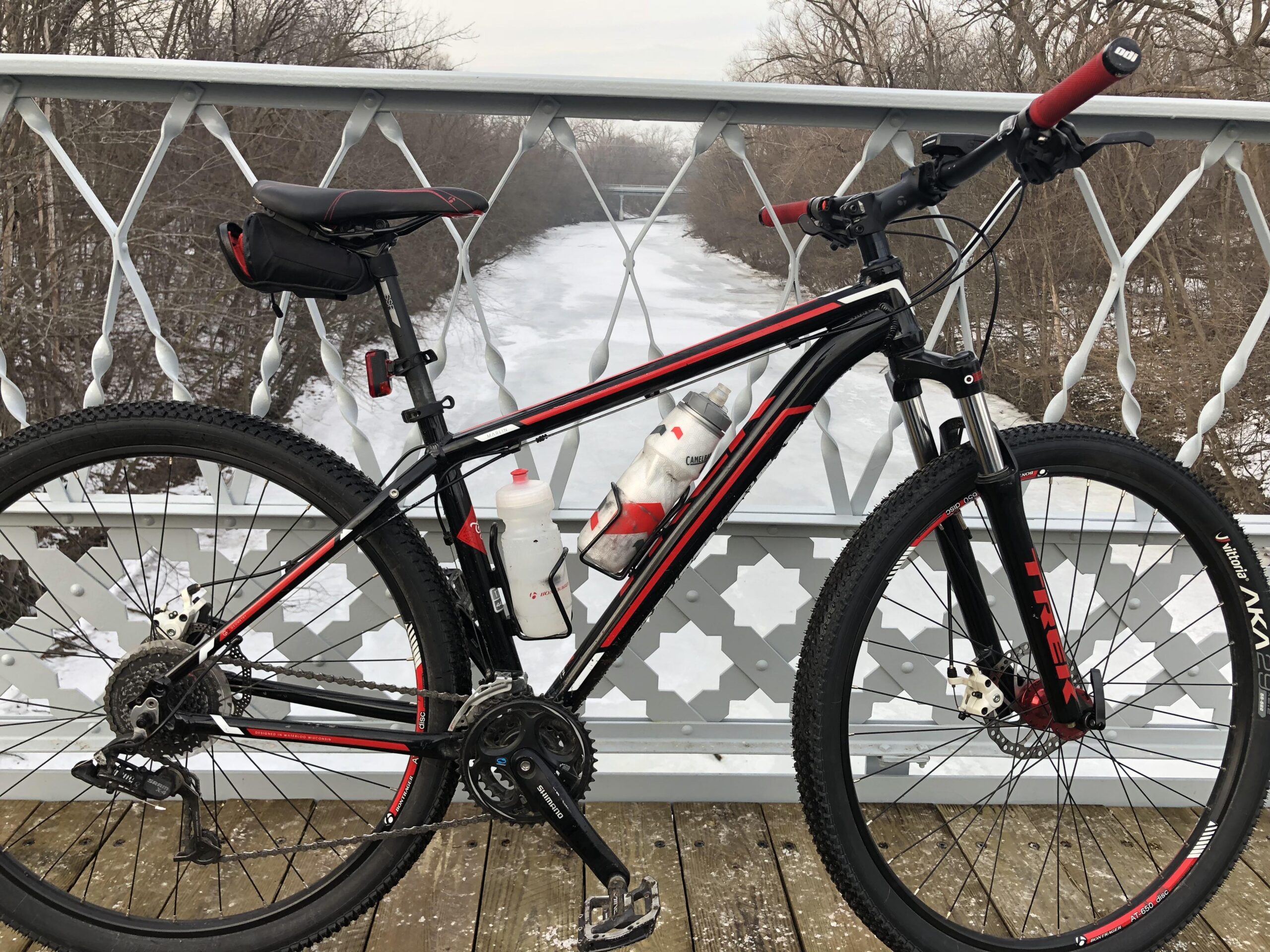 Trek Marlin: A black and red mountain bike is positioned on a wooden bridge, with a frozen river and trees visible in the background. The bike has two water bottles attached and is resting against a decorative railing. The scene conveys a serene outdoor winter atmosphere.