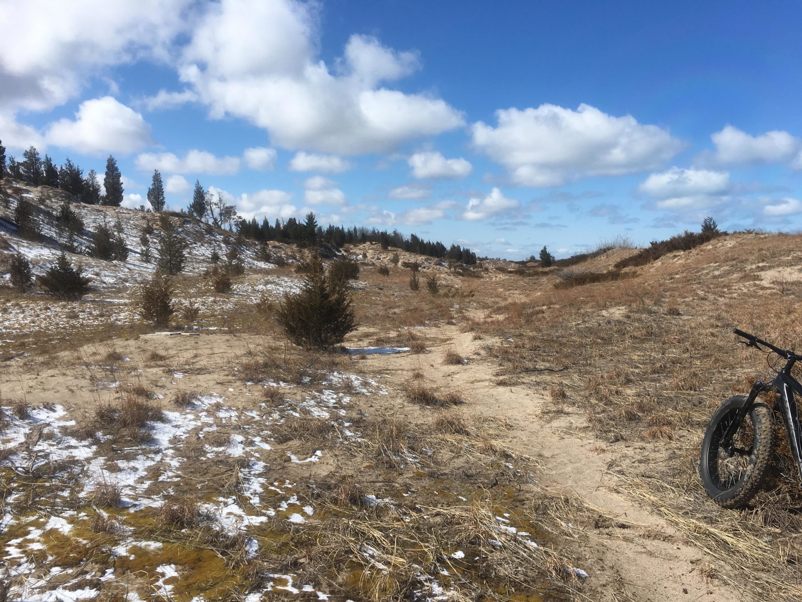 A scenic outdoor landscape featuring a sandy, dry terrain with patches of snow and sparse vegetation. In the foreground, a fat tire bike rests on the ground. The background displays rolling hills and a blue sky filled with fluffy, white clouds. Pinery Provincial Park mountain bike trail.