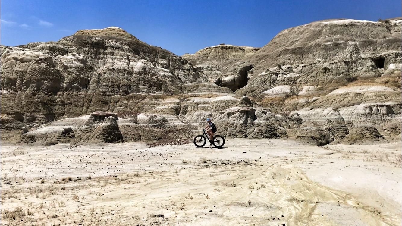 A cyclist riding a fat bike across a desert landscape with rugged, multi-colored rock formations under a clear blue sky. Double Slot Fat Bike Trail mountain bike trail.