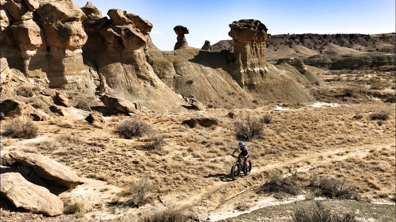 A mountain biker riding along a dirt trail in a rugged, desert landscape with unique rock formations and sparse vegetation under a clear blue sky. Double Slot Fat Bike Trail mountain bike trail.