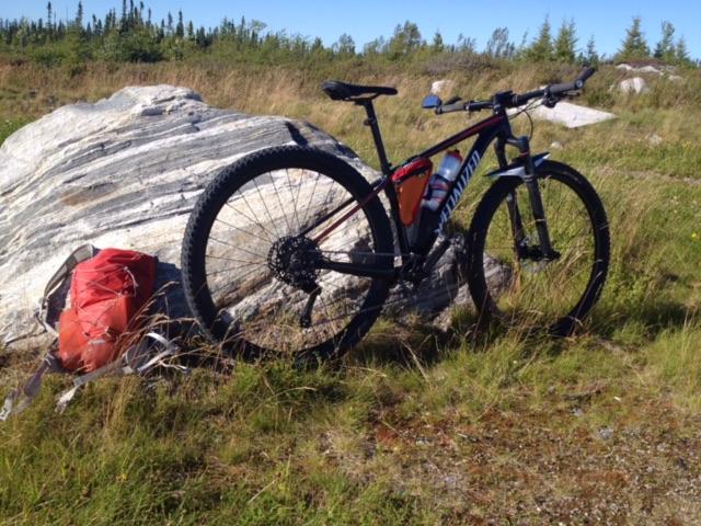 Specialized Epic Hardtail: A mountain bike leaning against a large rock in a grassy field, with a red backpack on the ground nearby. In the background, there are trees and open sky, suggesting an outdoor setting.