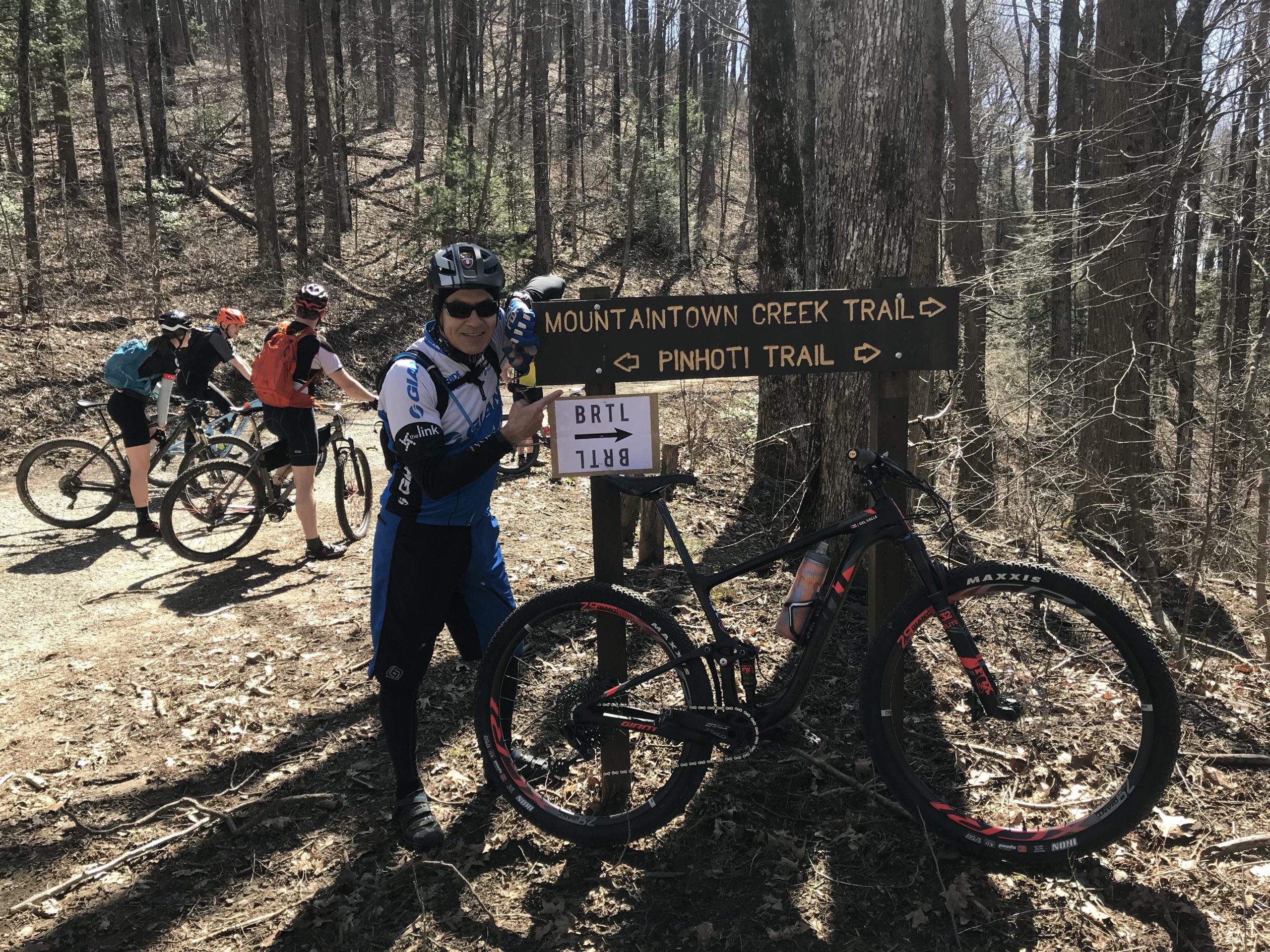 A group of mountain bikers paused at a trailhead sign indicating the Mountaintown Creek Trail and Pinhoti Trail. One cyclist stands in the foreground, smiling and pointing to the sign, while three others are seen in the background preparing their bikes. The scene is set in a wooded area with bare trees and fallen leaves on the ground, suggesting an early spring day. Pinhoti: Tatum Lead mountain bike trail.